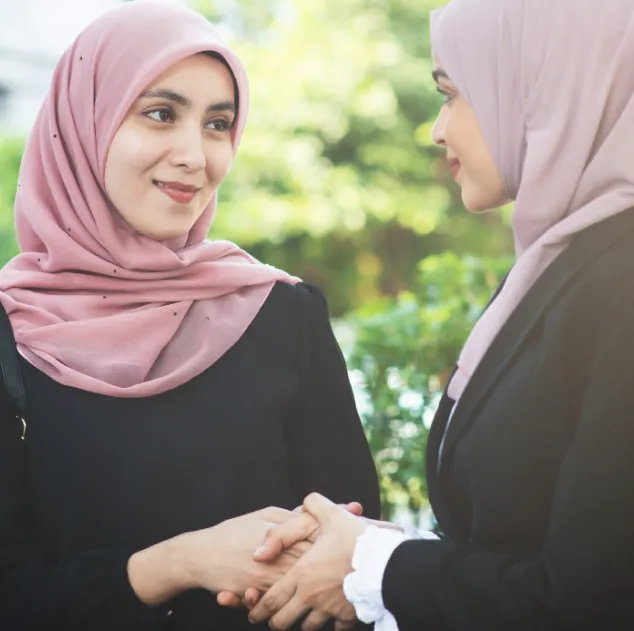 Two Muslim women wearing hijabs and professional attire smile while shaking hands outdoors against a soft-focus background of green foliage.