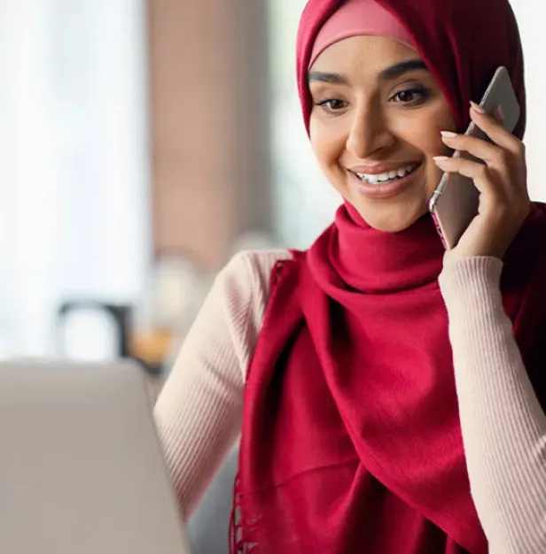 A smiling woman wearing a red hijab talks on her smartphone while looking down at a laptop screen.