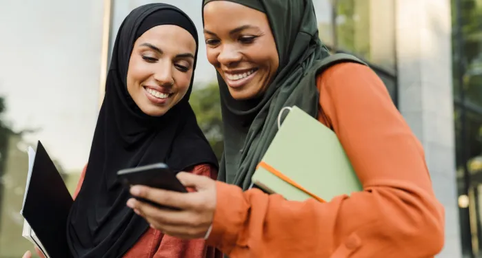 Two smiling women wearing hijabs stand together outdoors, looking down at a smartphone held by one of them while carrying notebooks.