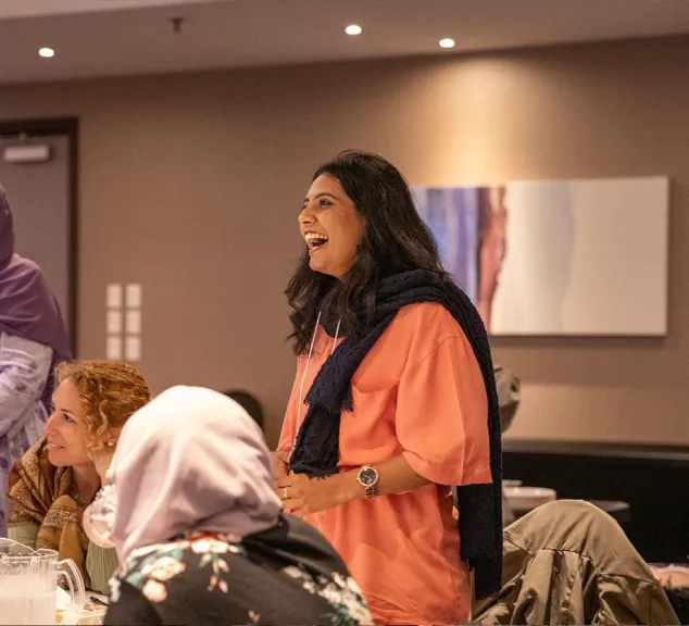 A woman with long dark hair, wearing a peach-colored top and a dark blue scarf, laughs while standing in a room with other people seated around her.