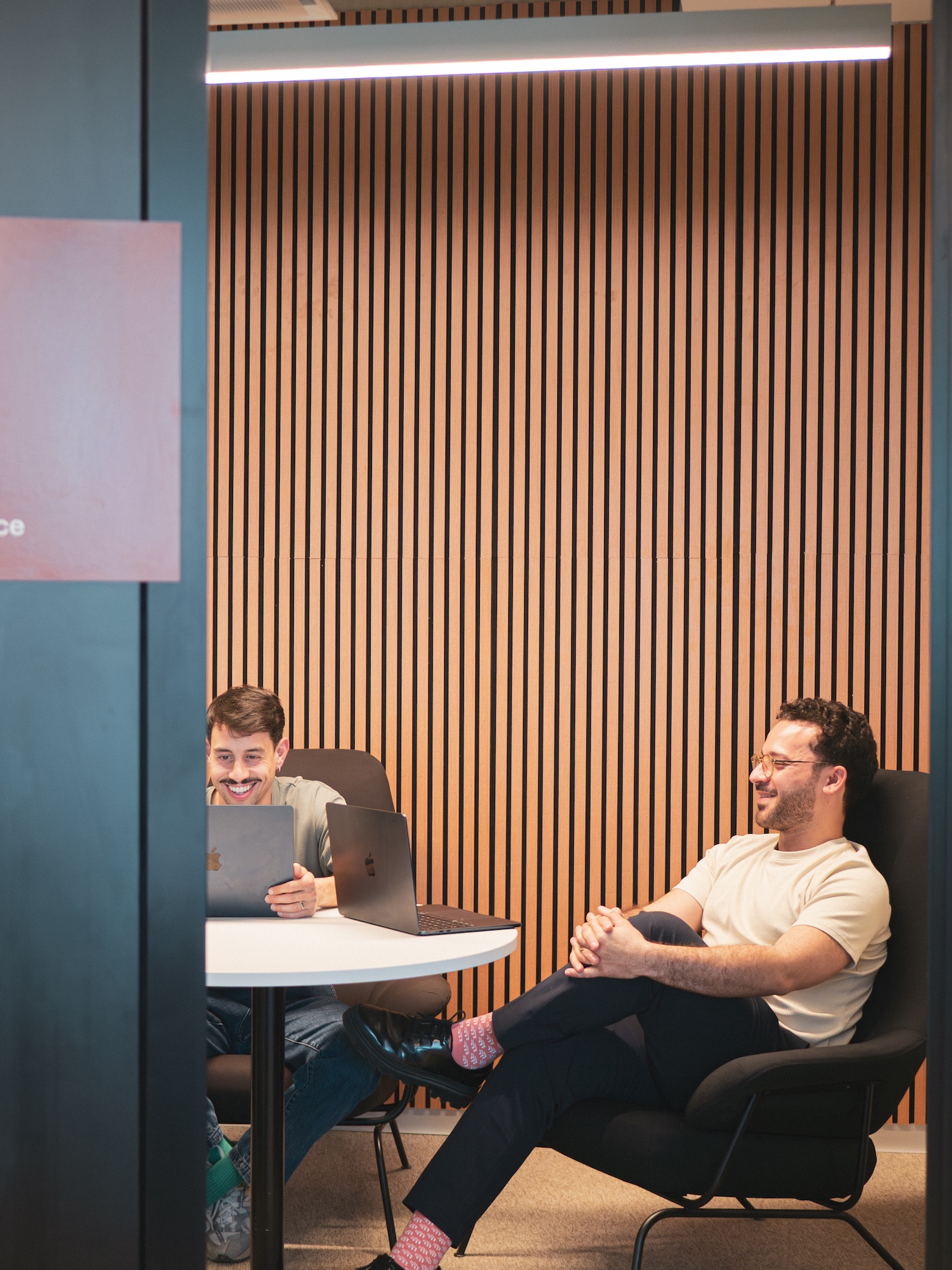 Two Hive colleagues laughing over laptops at a round table in a wood-slatted meeting room