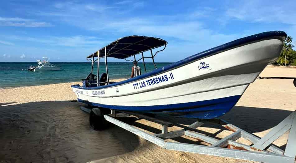 White and blue motorboat named 'TTT Las Terrenas II' parked on sandy beach with a blue canopy and calm ocean in the background under a partly cloudy sky.
