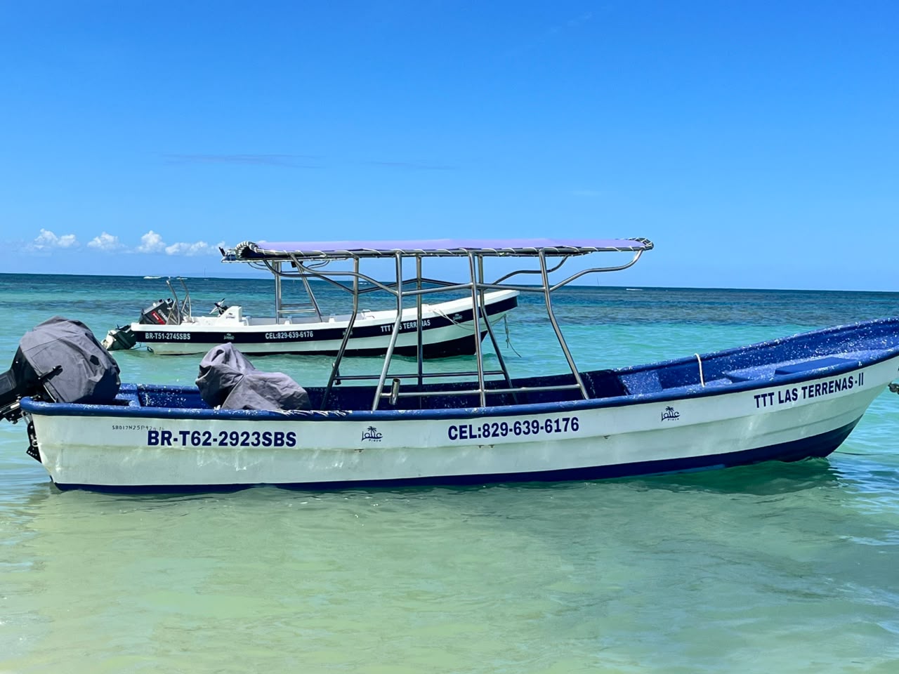 Two small motorboats floating in clear shallow turquoise water under a bright blue sky, with visible registration numbers and a phone number on the boats' sides.