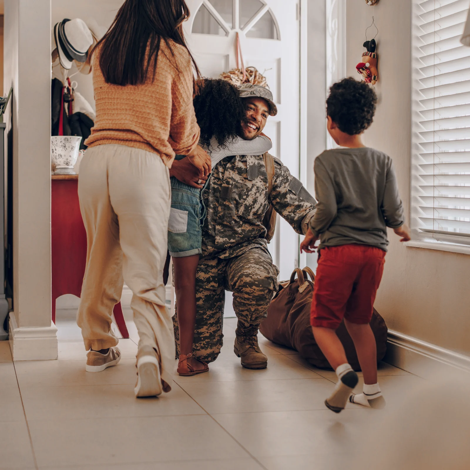 A soldier in camouflage uniform kneeling and smiling while hugging a child as a woman and another child approach inside a home.