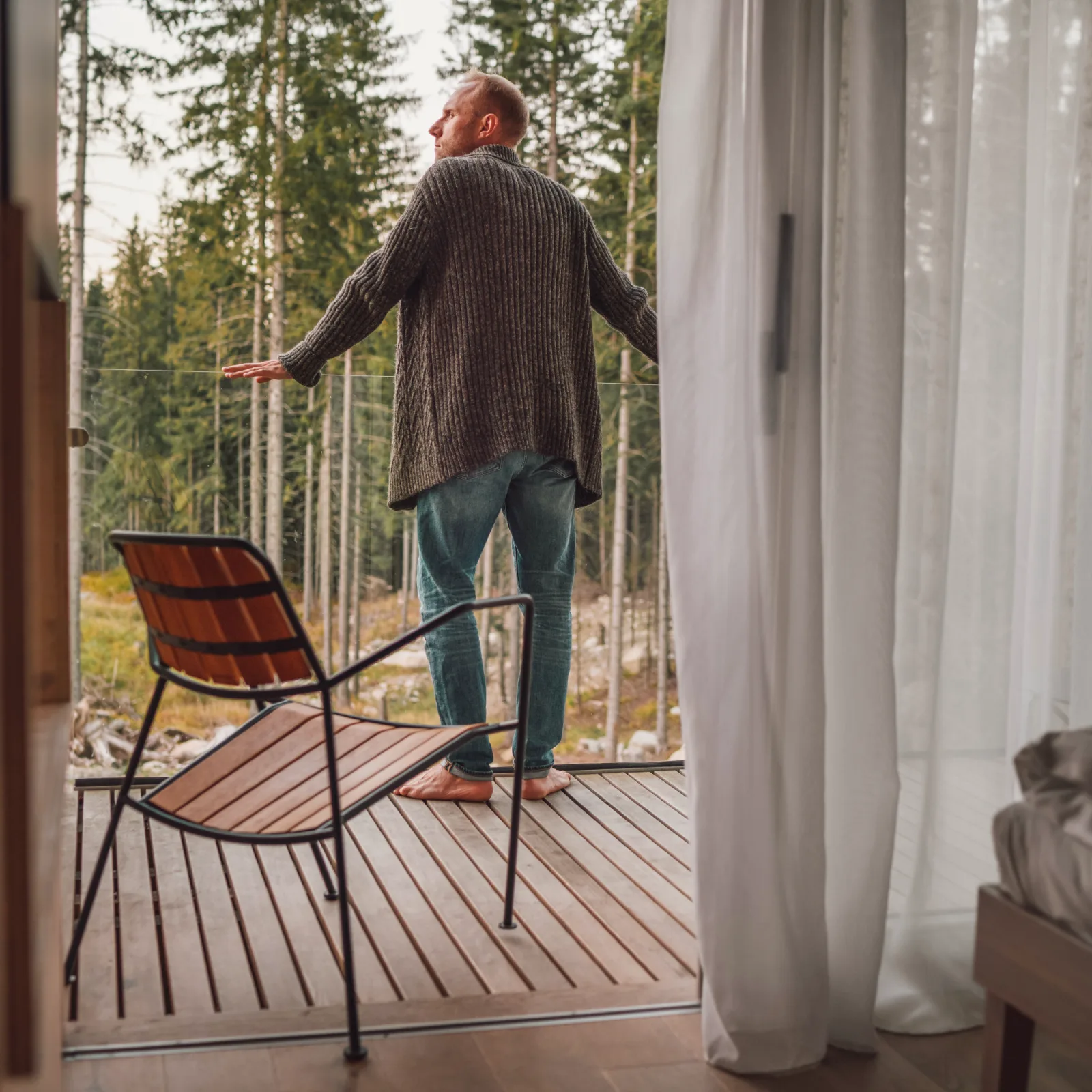 Man in a gray sweater and jeans standing barefoot on a wooden balcony overlooking a forest.