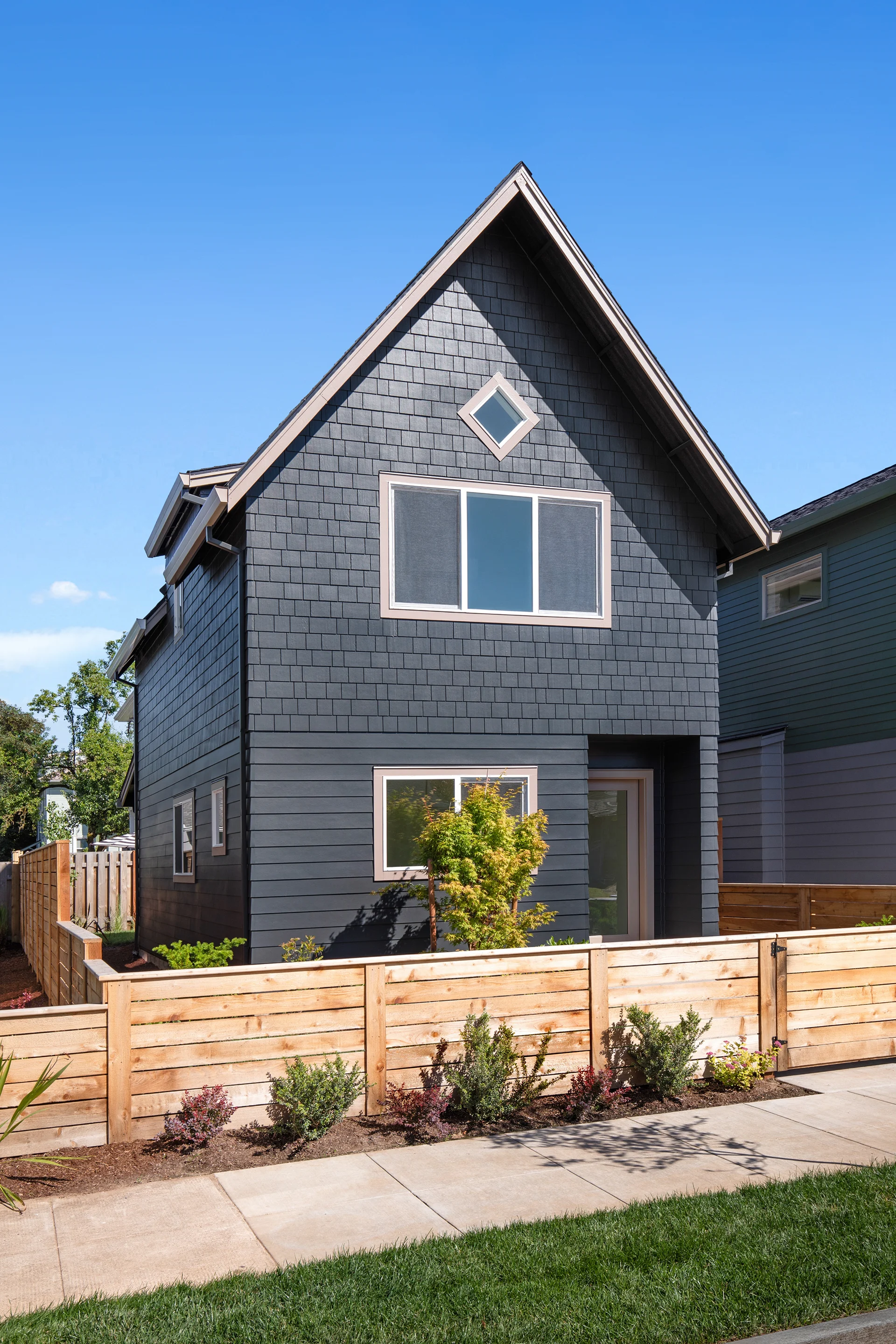 Two-story dark gray house with shingle siding, wooden fence, and small bushes in front under clear blue sky.