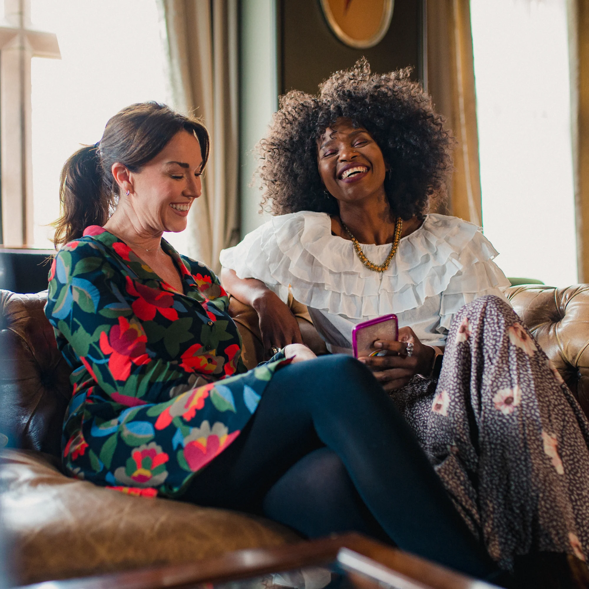 Two women sitting on a leather couch, smiling and enjoying a conversation, one holding a phone.