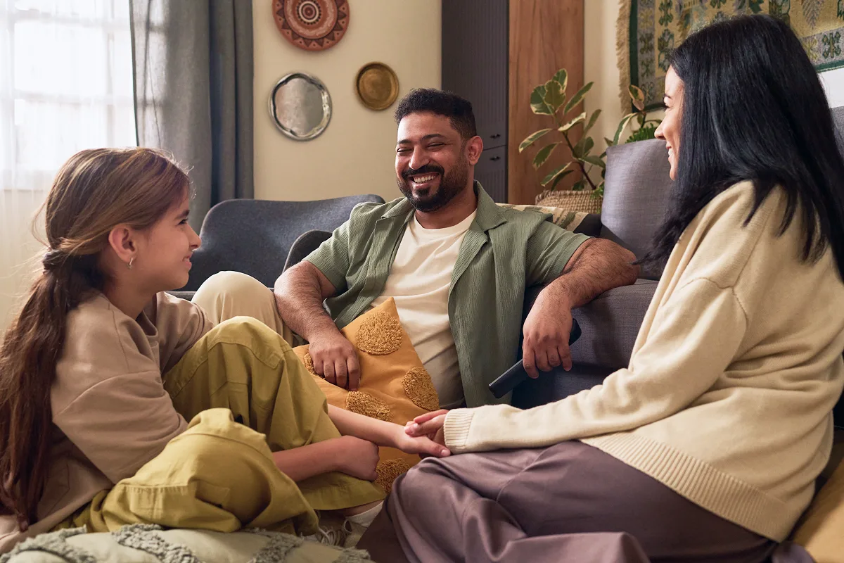Smiling family of three sitting on the floor and couch in a cozy living room, holding hands and enjoying time together.