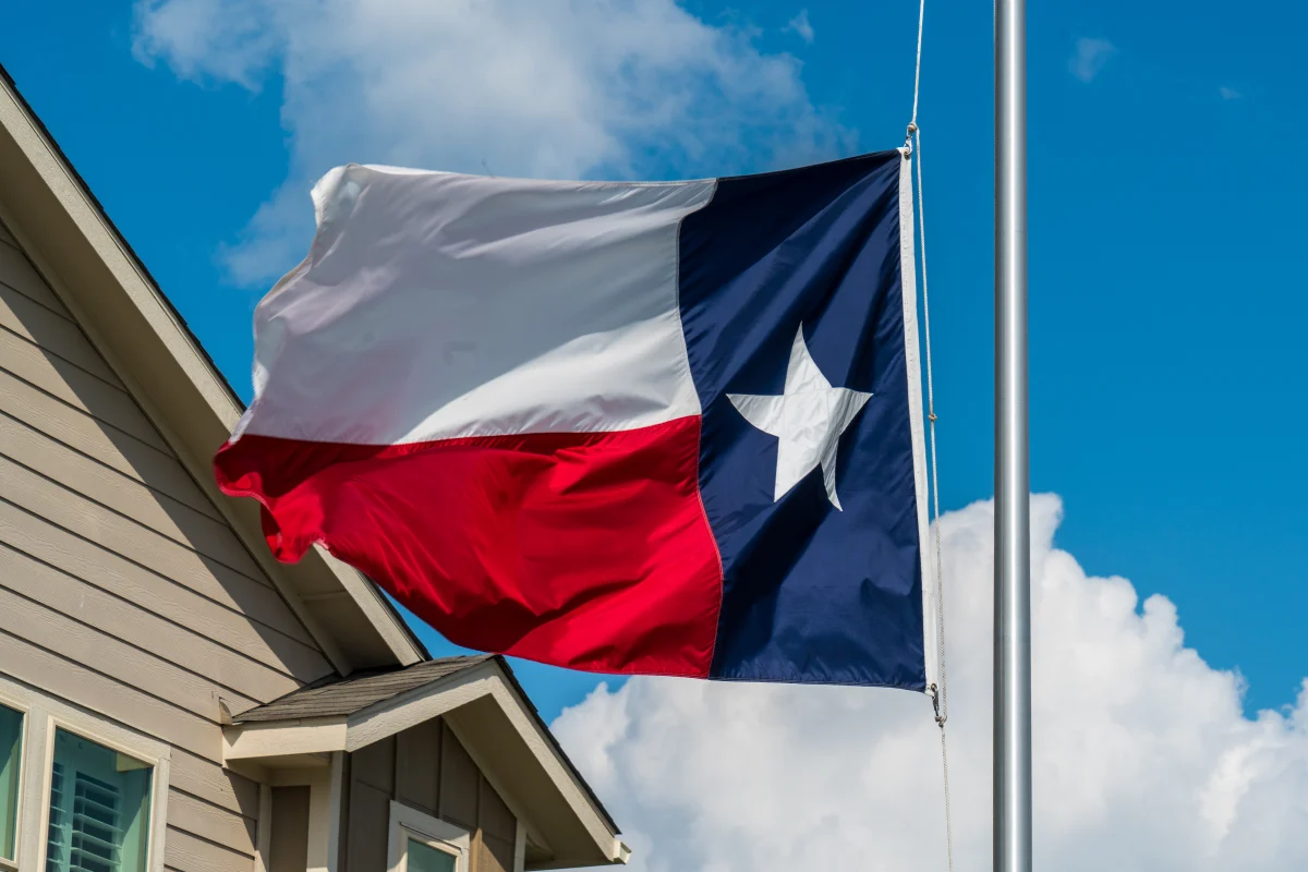 Texas state flag waving on a flagpole next to a house roof under a blue sky with clouds.