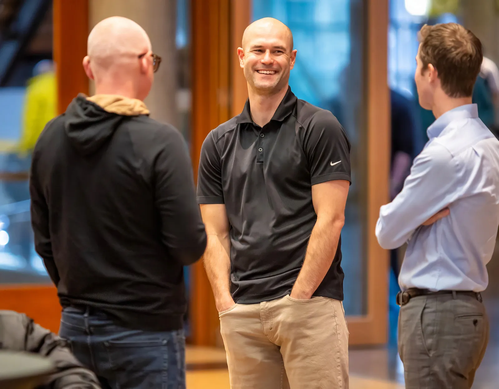 Three men in casual and business casual attire engaged in conversation indoors, one smiling with hands in pockets.