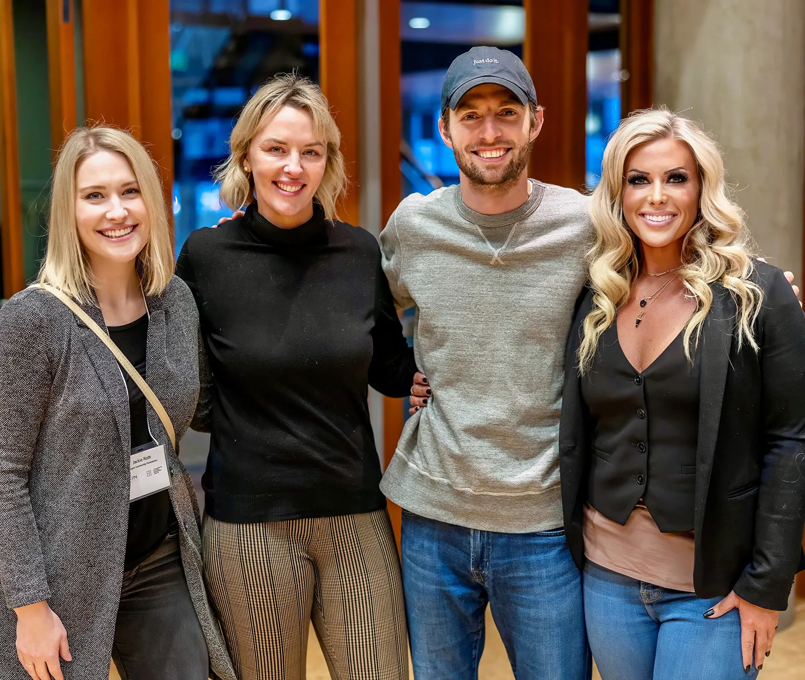 Four smiling adults standing closely together indoors, three women and one man wearing casual and semi-formal clothing.