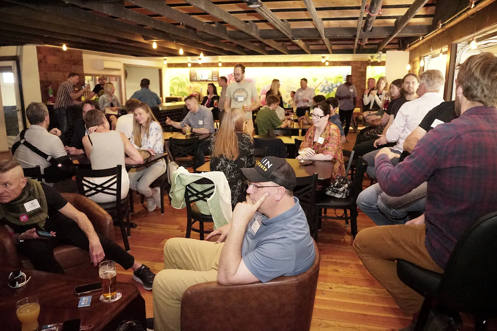 A lively indoor gathering with people chatting, sitting at tables and chairs, enjoying drinks in a warmly lit room with wooden floors and exposed beams.