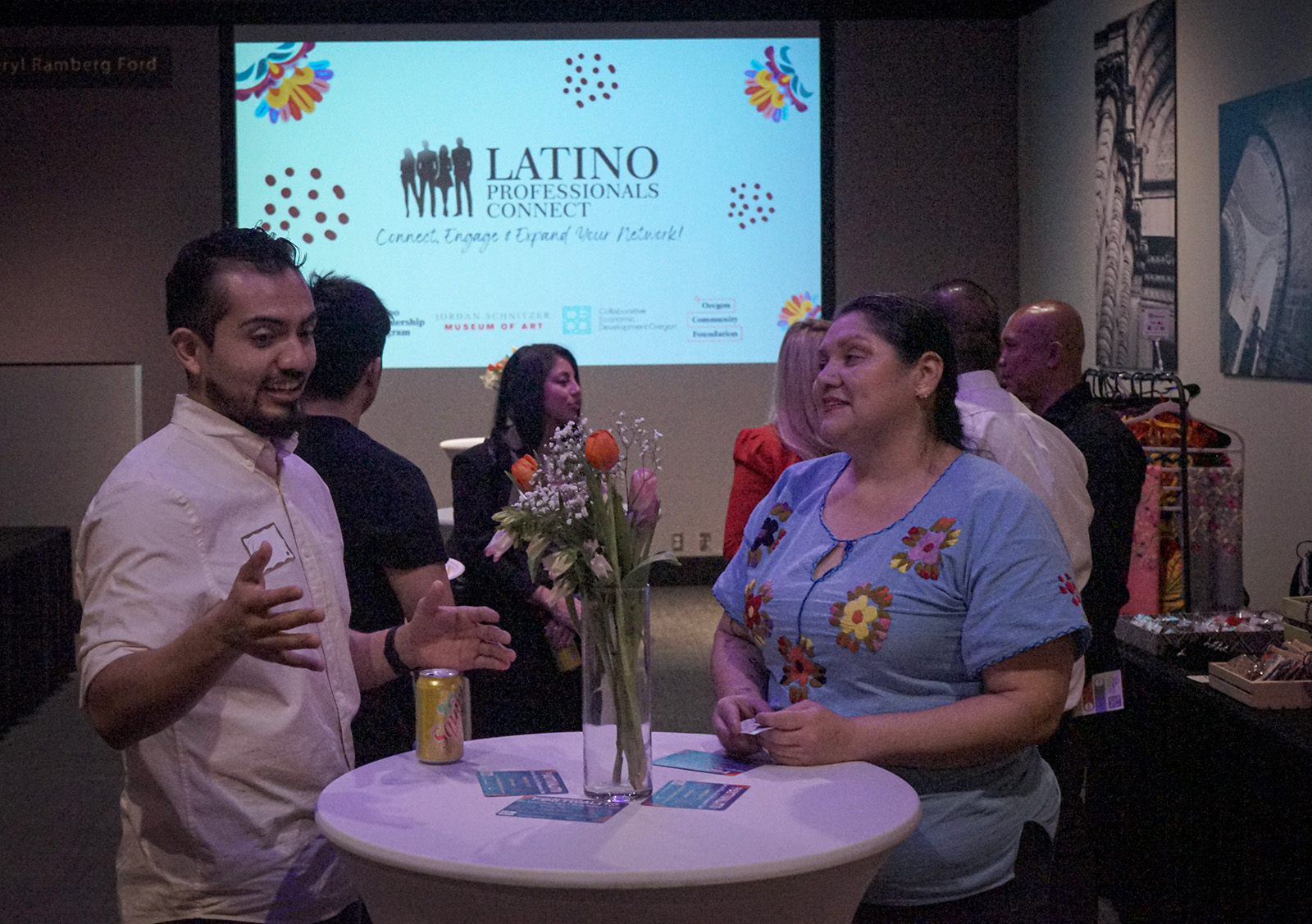 Two people engaging in conversation at a networking event with a sign reading Latino Professionals Connect in the background.