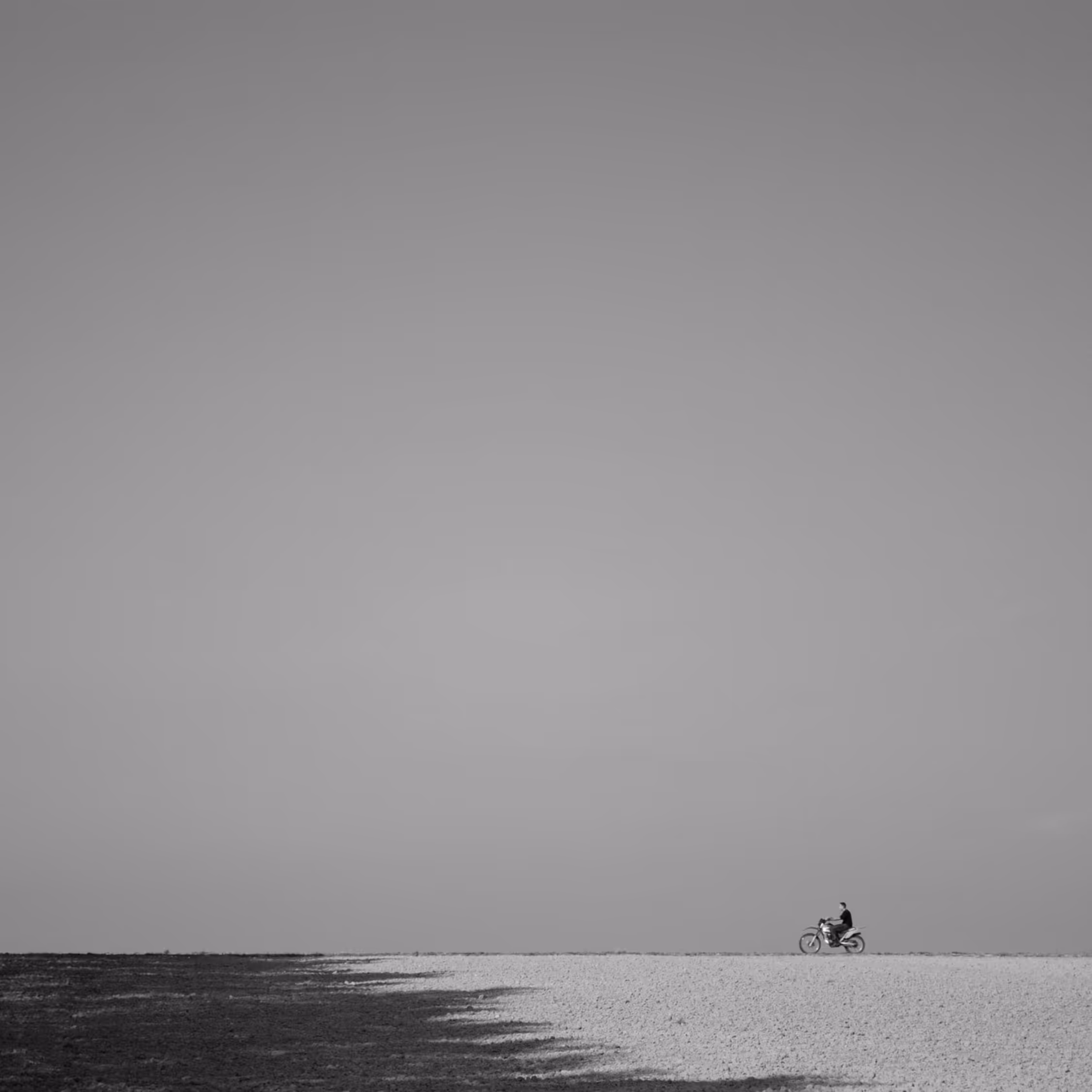 Minimalist black and white fine art photography featuring a tiny solitary motorcyclist riding along the horizon line against a vast, empty sky, emphasizing negative space and solitude.