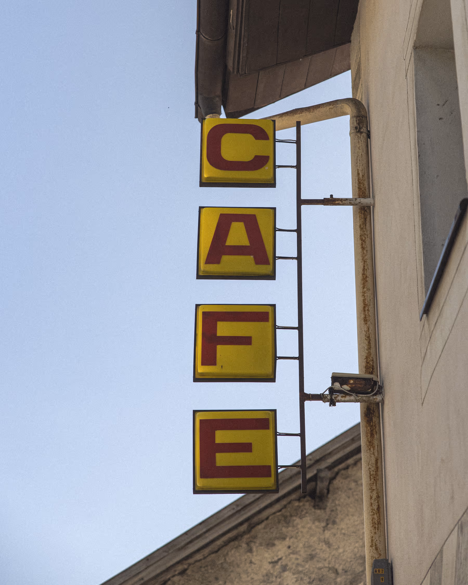 Minimalist travel photography from South Tyrol, Italy. A vintage yellow vertical 'CAFE' sign on a beige facade featuring visible film grain and graphic composition.