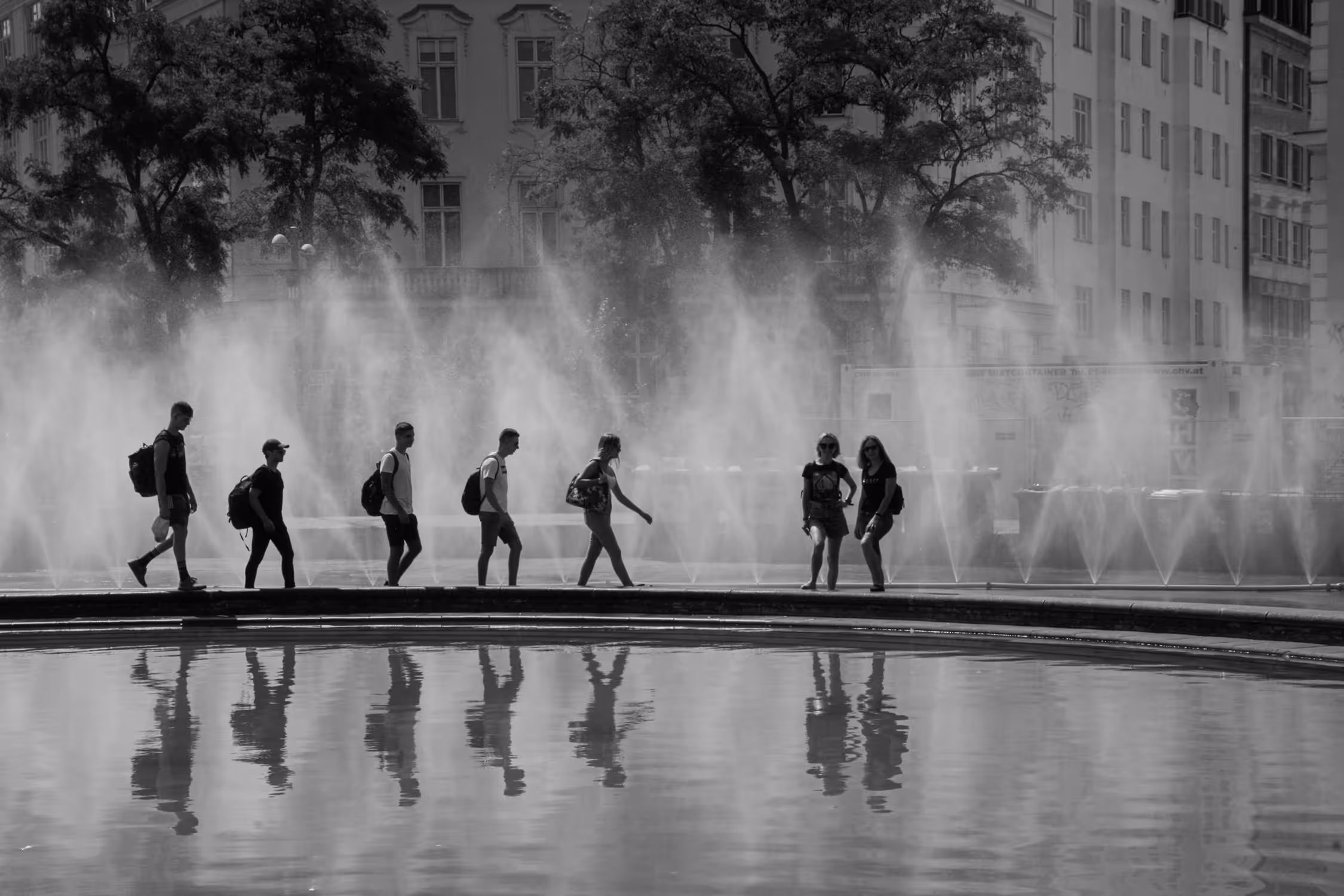 Atmospheric black and white street photography at Schwarzenbergplatz, Vienna, capturing silhouettes of people against the mist of the Hochstrahlbrunnen fountain, mirrored by a sharp reflection in the foreground water.