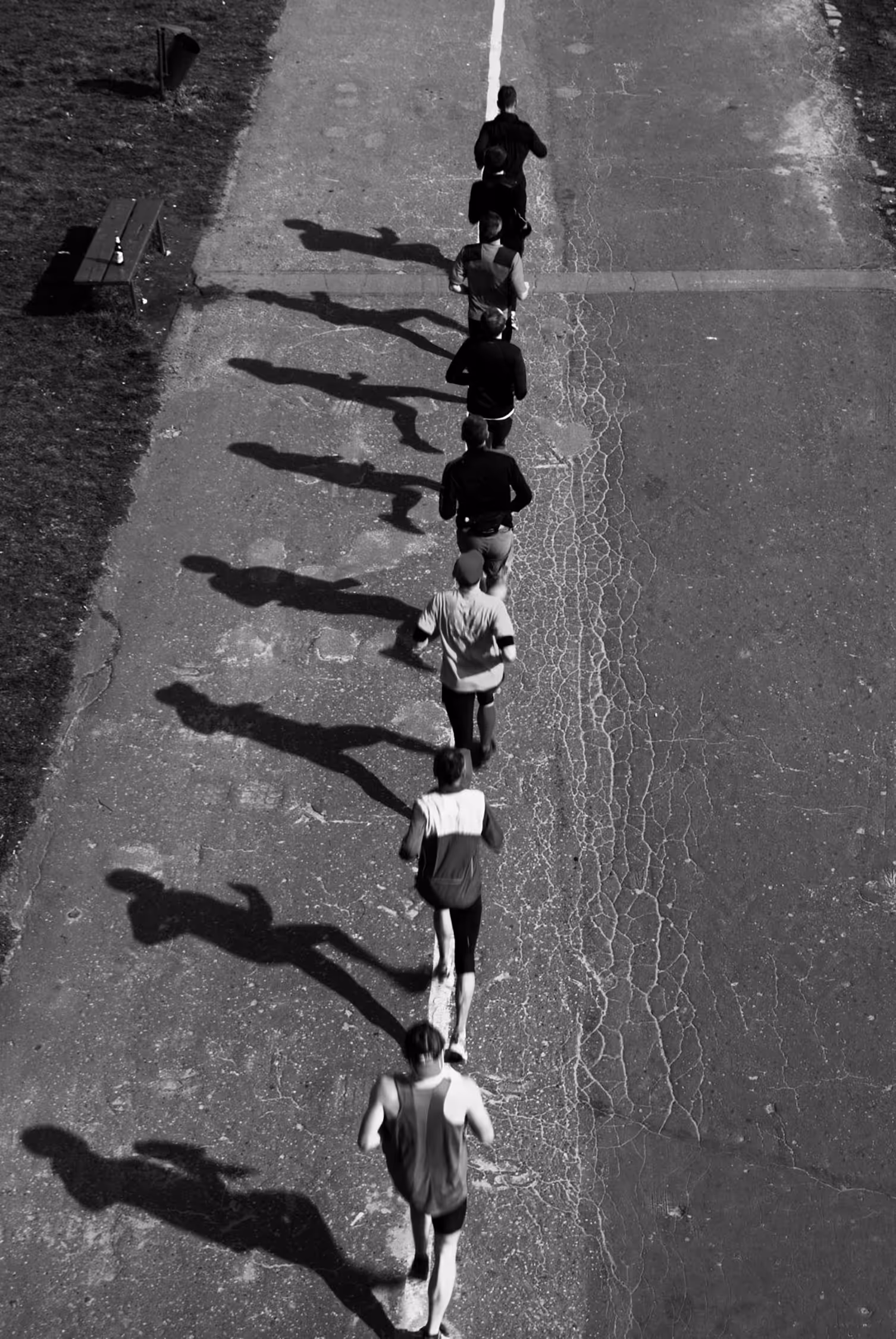 High-angle black and white street photography in Krakow featuring a line of runners on pavement. Harsh sunlight creates long, dramatic shadows that form a rhythmic, geometric pattern mirroring the athletes' movement.