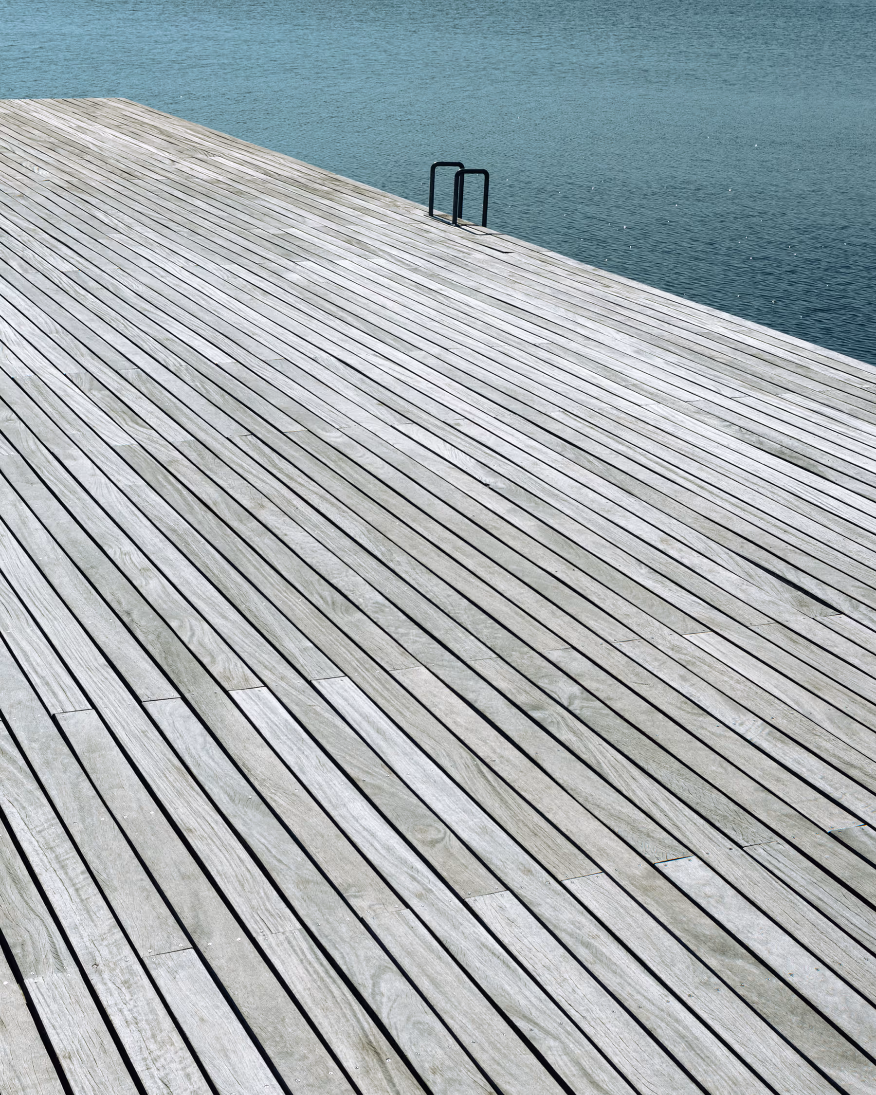 Minimalist architectural photography of the wooden promenade near The Wave (Bølgen) building in Vejle, featuring strong diagonal lines of timber decking contrasting with deep teal water.