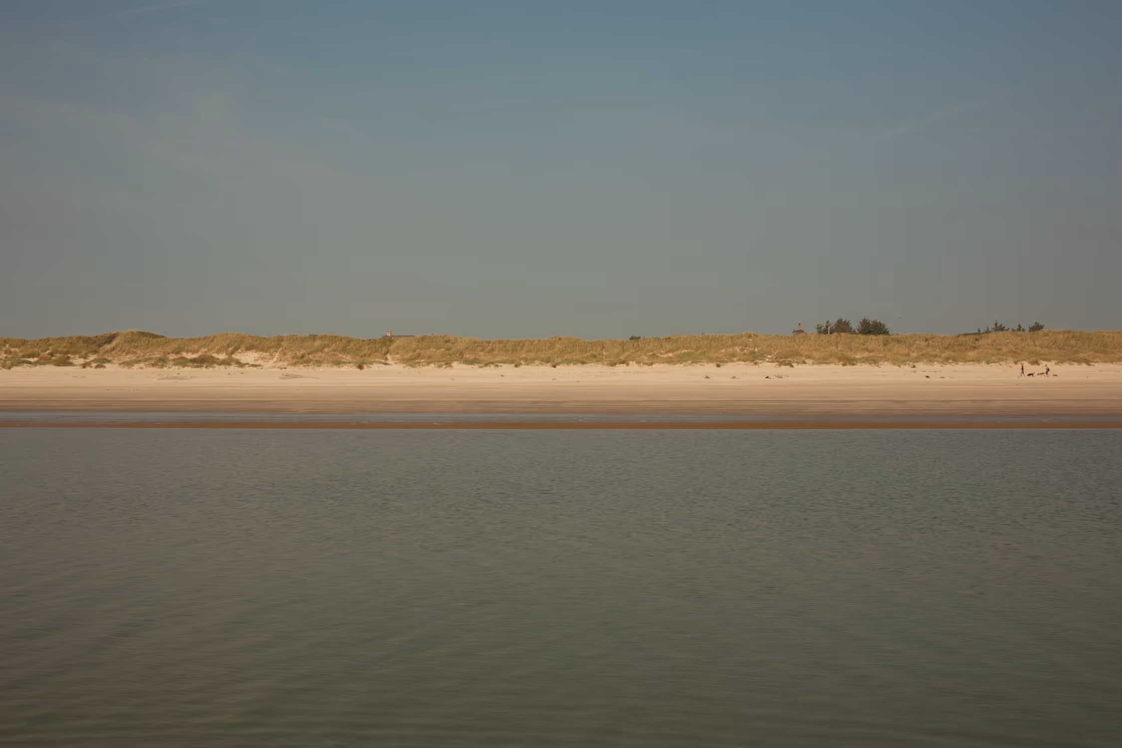 A serene, minimalist seascape of Blavand beach in Denmark, featuring distinctive horizontal layers of calm water, sandy shore, and grassy dunes set against a vast, clear sky with tiny figures emphasizing the scale.