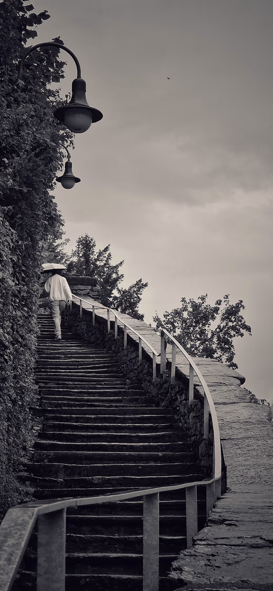 Atmospheric black and white vertical shot of stone stairs leading to Burgruine Ringgenberg in Interlaken, featuring a lone figure with an umbrella and a vintage street lamp framing the composition with strong leading lines.