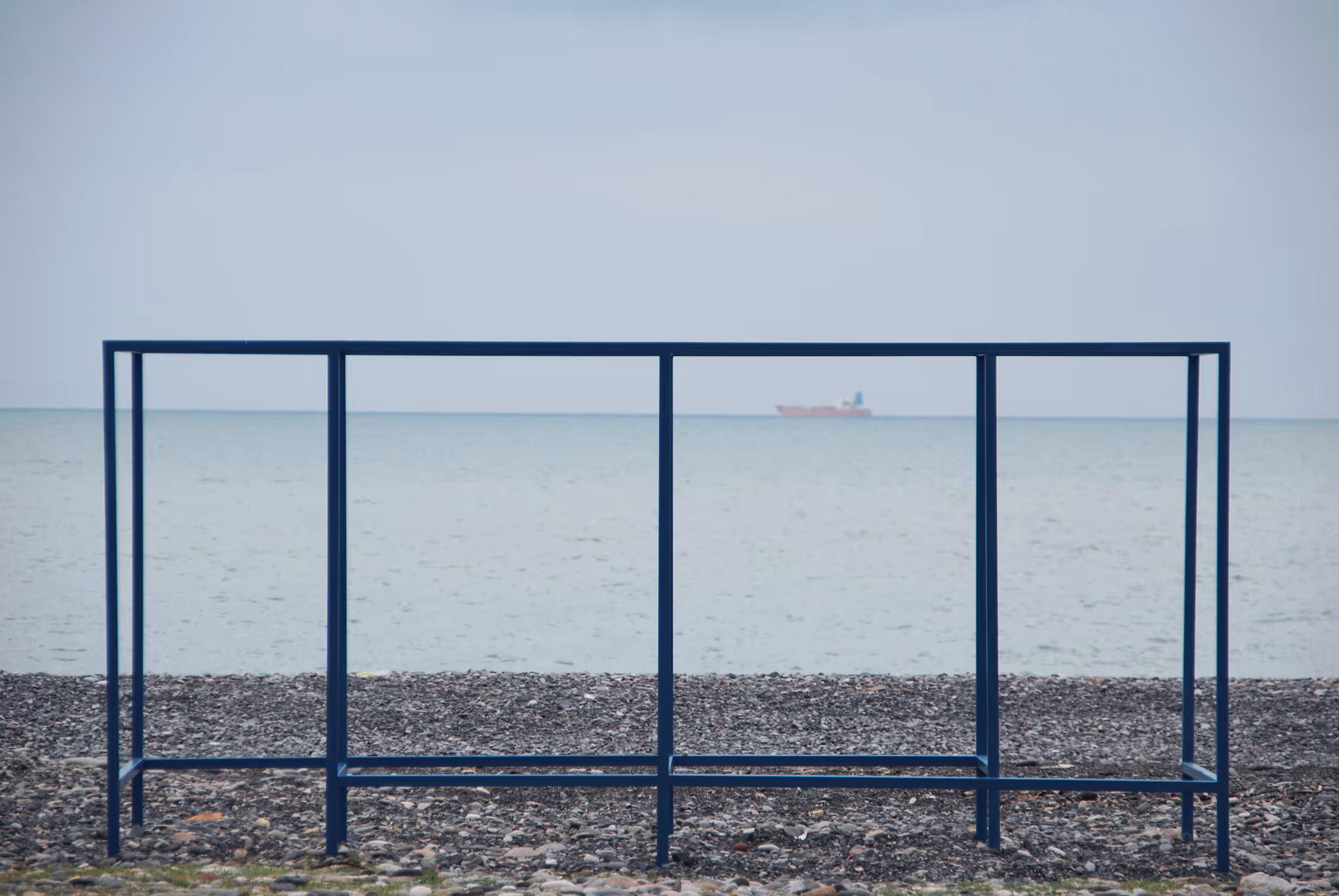 A minimalist seascape from Batumi beach featuring a blue rectangular metal frame standing on pebbles, creating a geometric composition against the calm horizon and a distant ship.