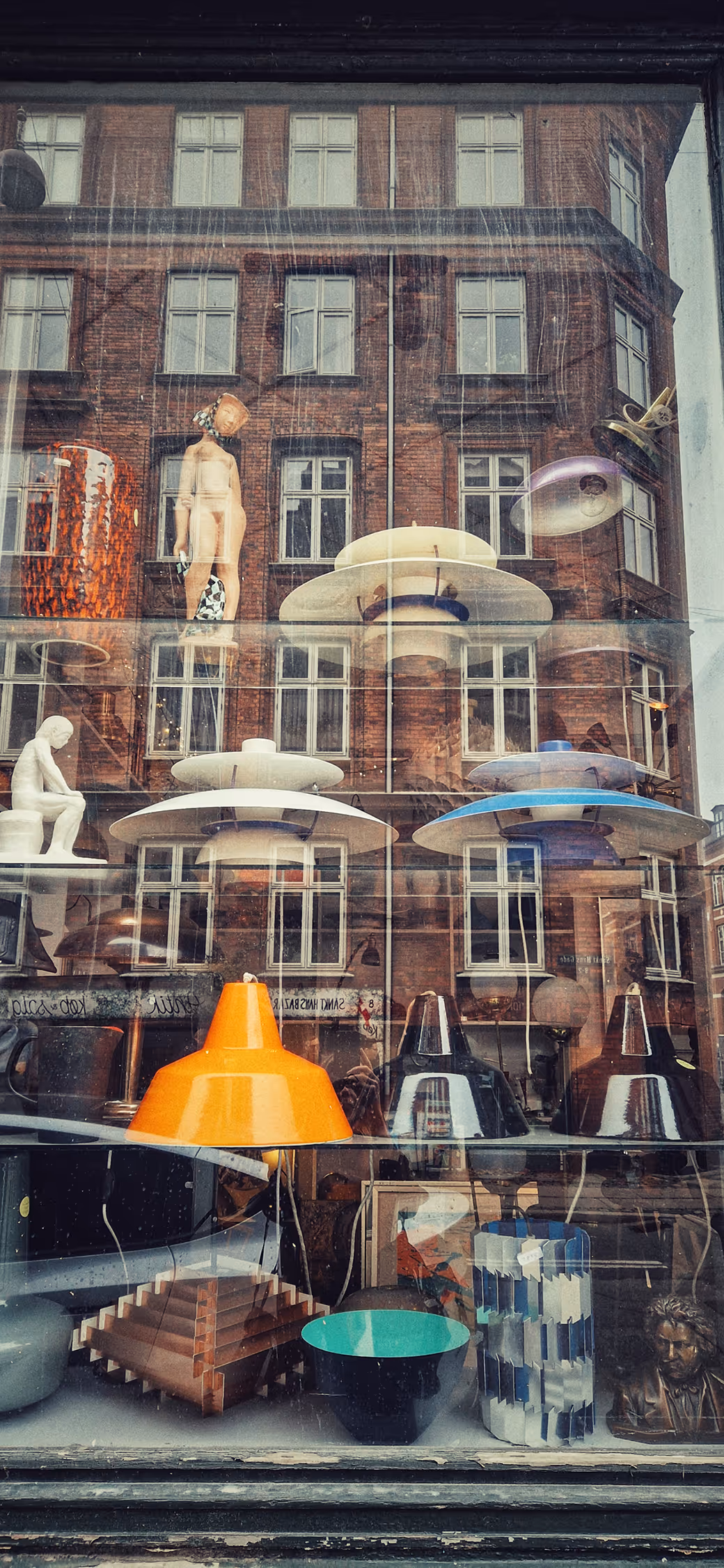 Urban photography of a vintage shop window in Copenhagen, visually layering iconic Danish design lamps (PH style) with a sharp reflection of a historic red brick facade to create a textured collage.