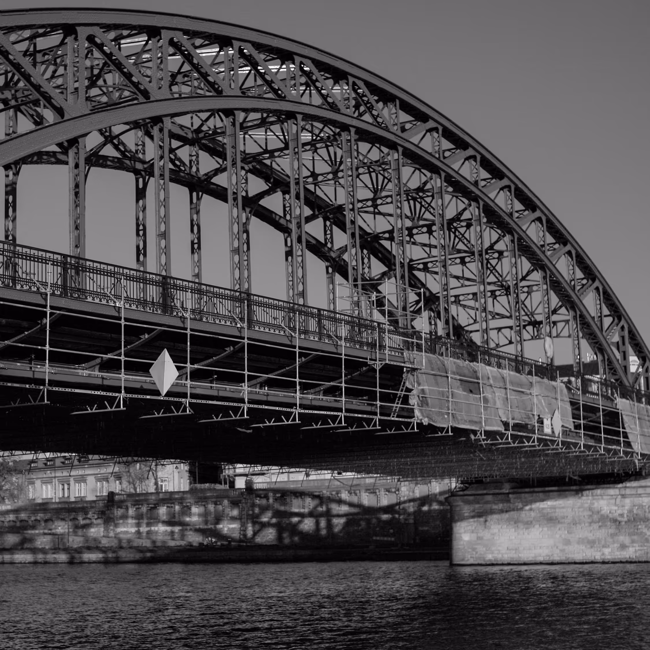 A dramatic black and white architectural shot of the Marshal Józef Piłsudski Bridge in Kraków, highlighting the heavy steel truss arch and raw industrial construction spanning over the Vistula River.