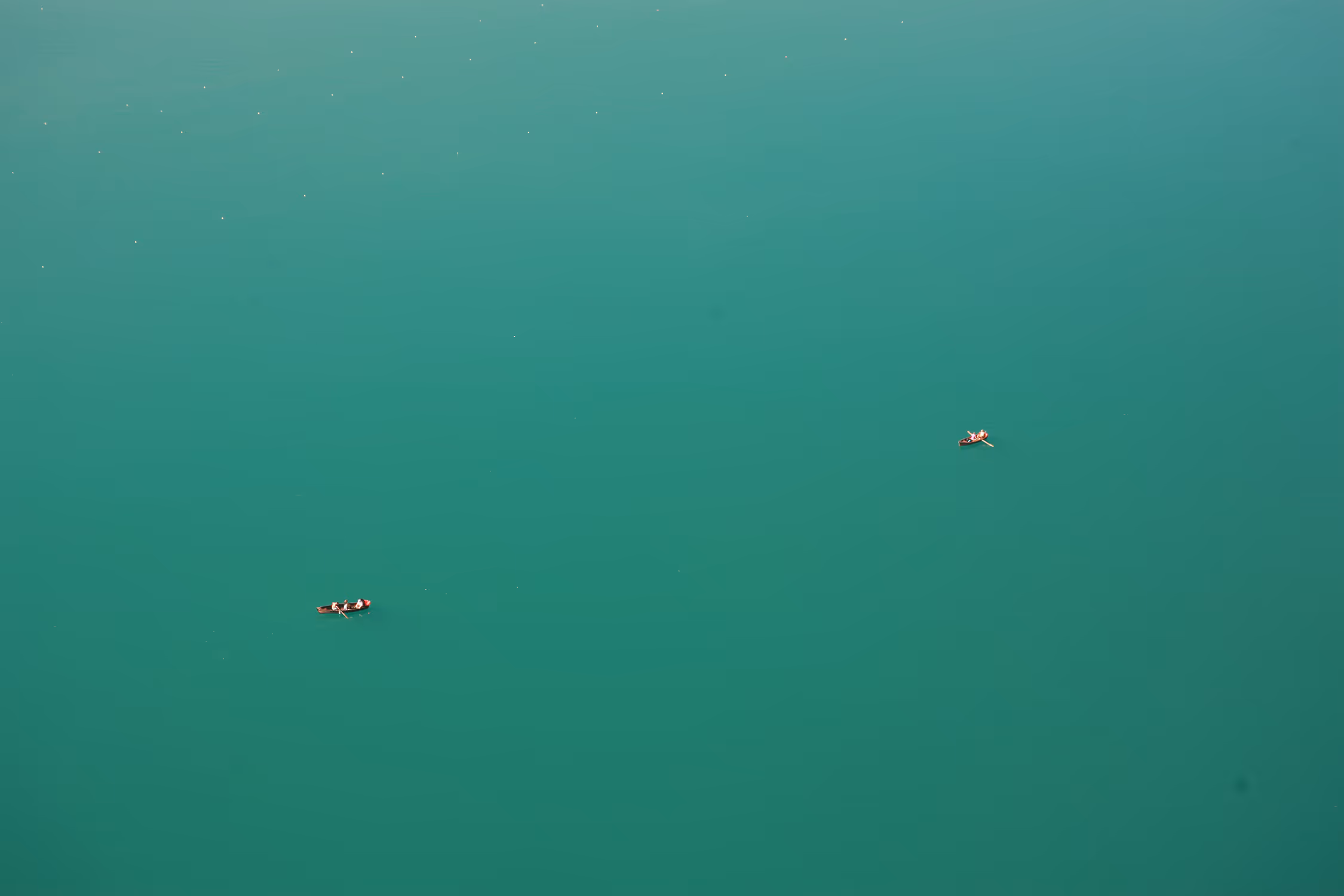 Minimalist high-angle view of Lake Bled in Slovenia, capturing tiny wooden rowboats floating in vast negative space of calm turquoise water, creating a serene and meditative atmosphere.