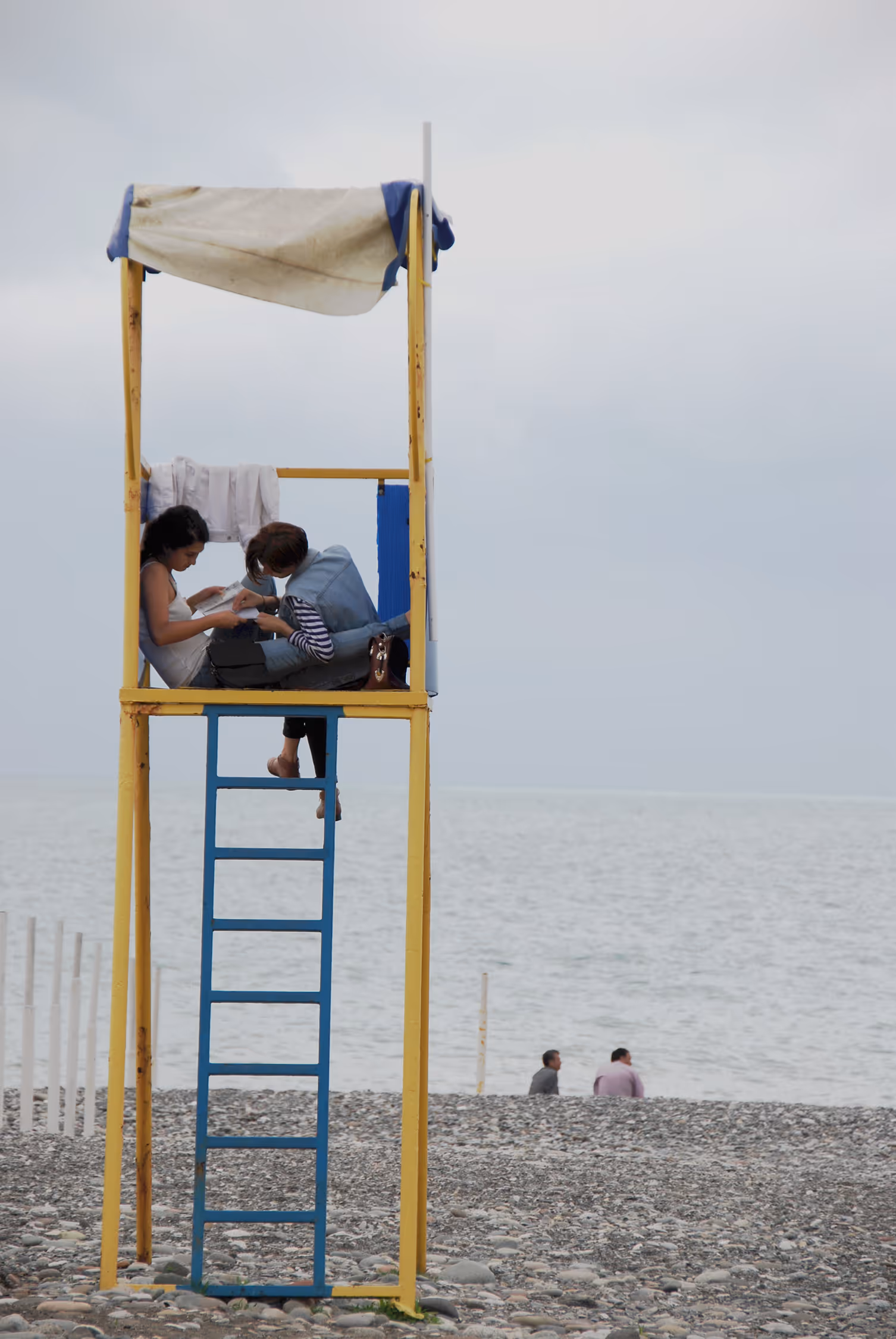 Candid lifestyle photograph of two women relaxing on a yellow lifeguard tower on a pebble beach in Batumi, Georgia, framed by negative space and a moody overcast sky.