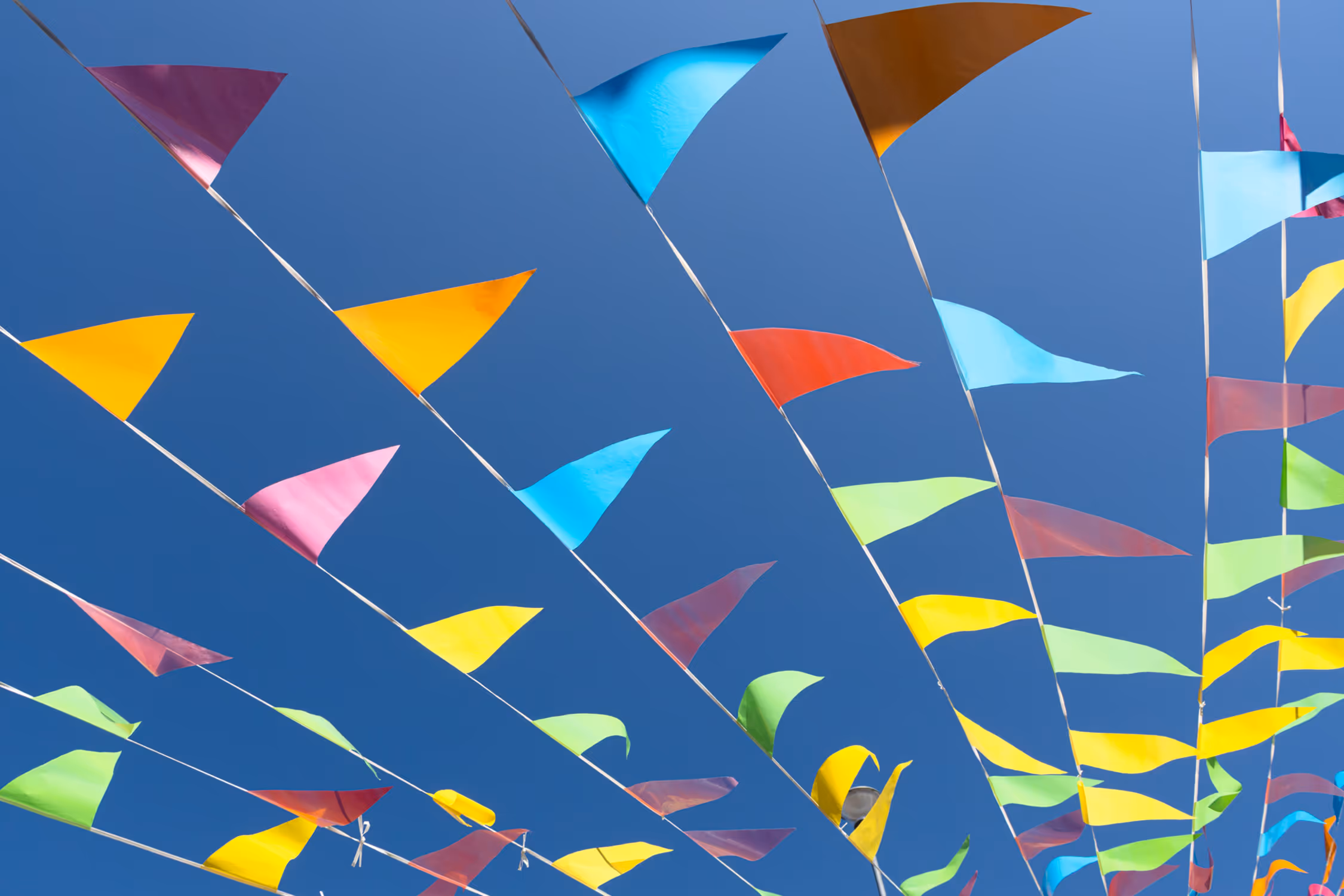 Low-angle shot of vivid triangular bunting flags against a deep blue sky in Vejle, creating a rhythmic diagonal pattern full of negative space and festive energy.