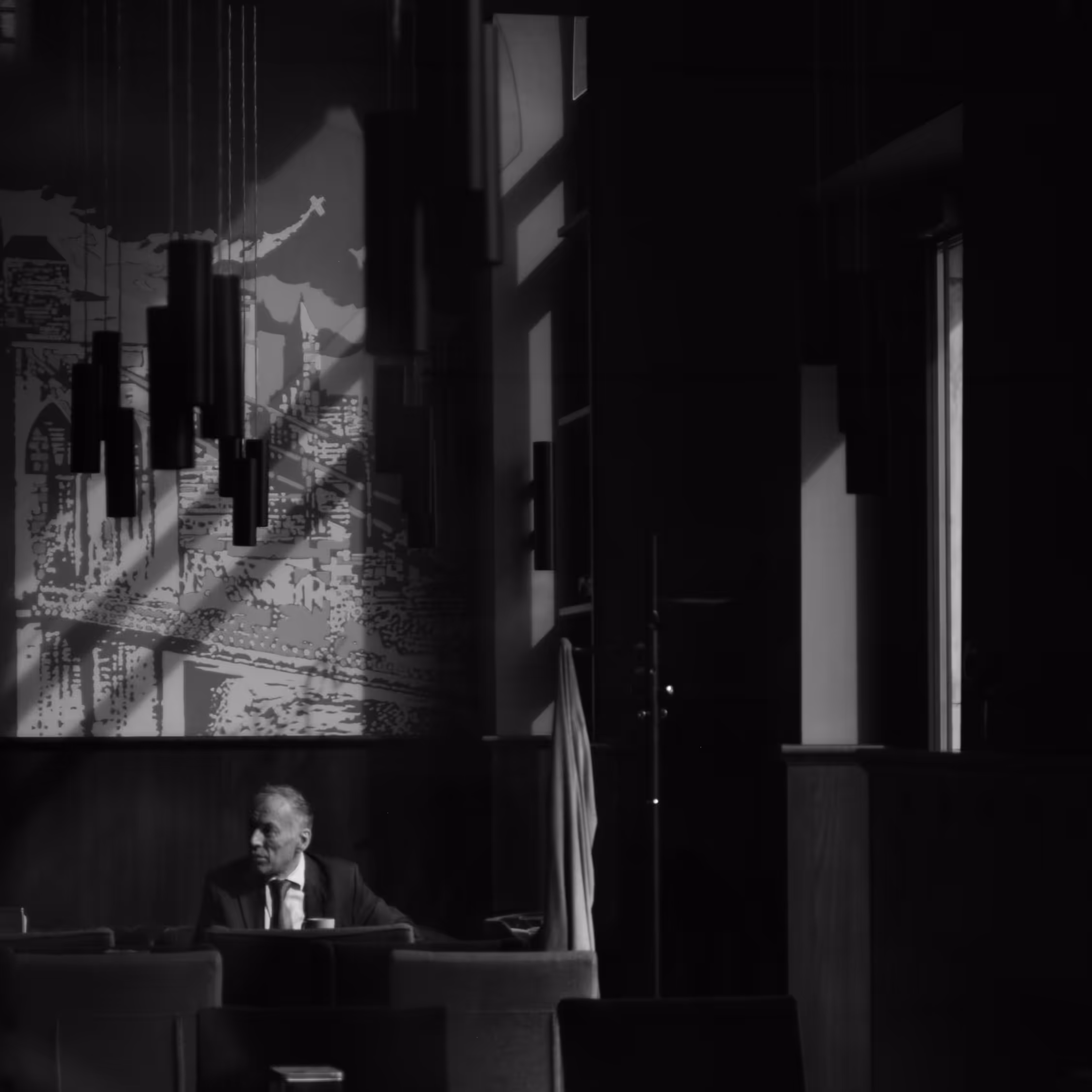 High-contrast monochrome candid portrait of a mature man in a suit seated alone in a dark Wrocław cafe, dramatically lit by window light creating sharp diagonal shadows and geometric shapes on the wall mural.