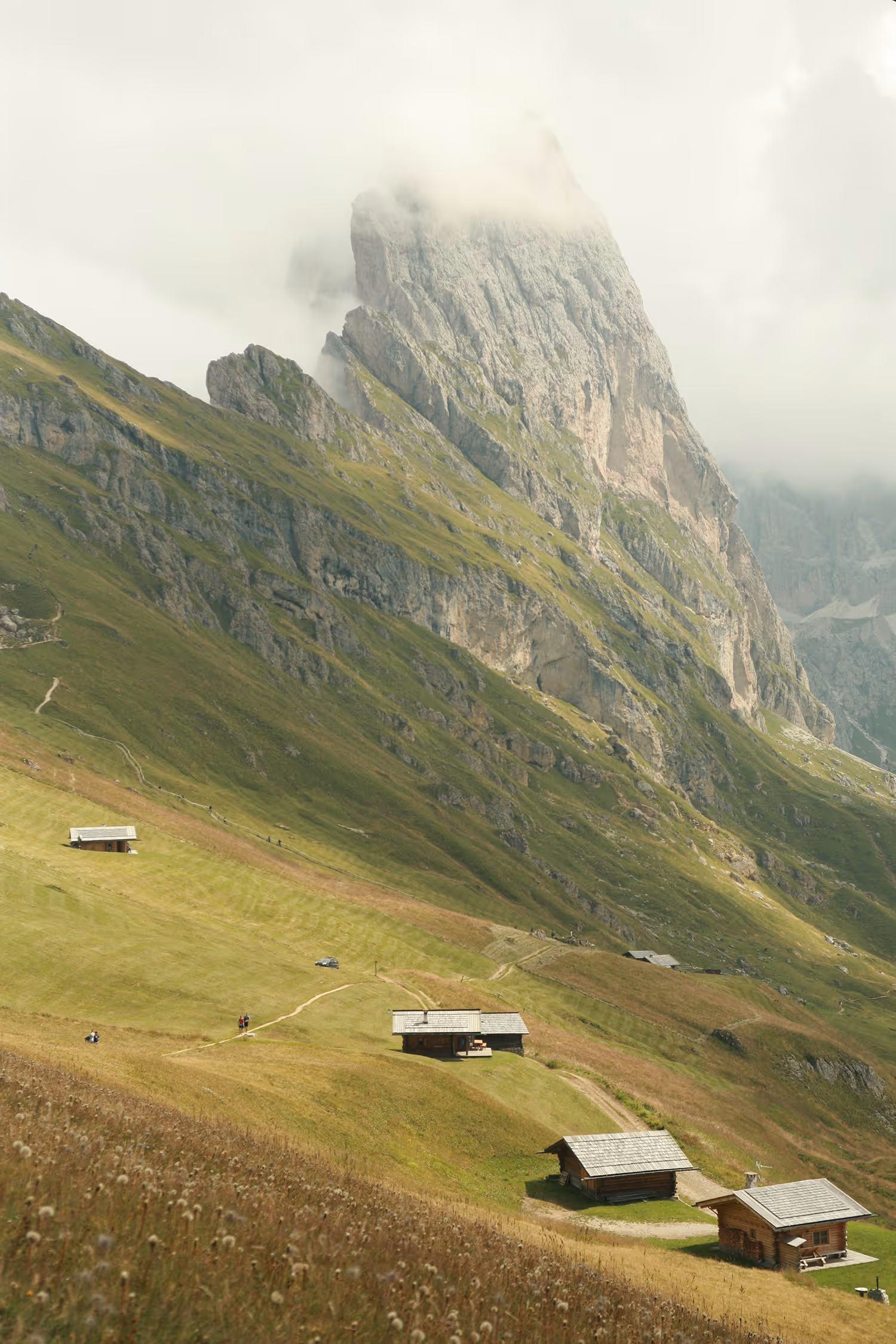Vertical landscape photograph of Seceda peak, emphasizing dramatic texture of the rock face against soft, green meadows with wooden cabins and leading lines of the hiking paths.