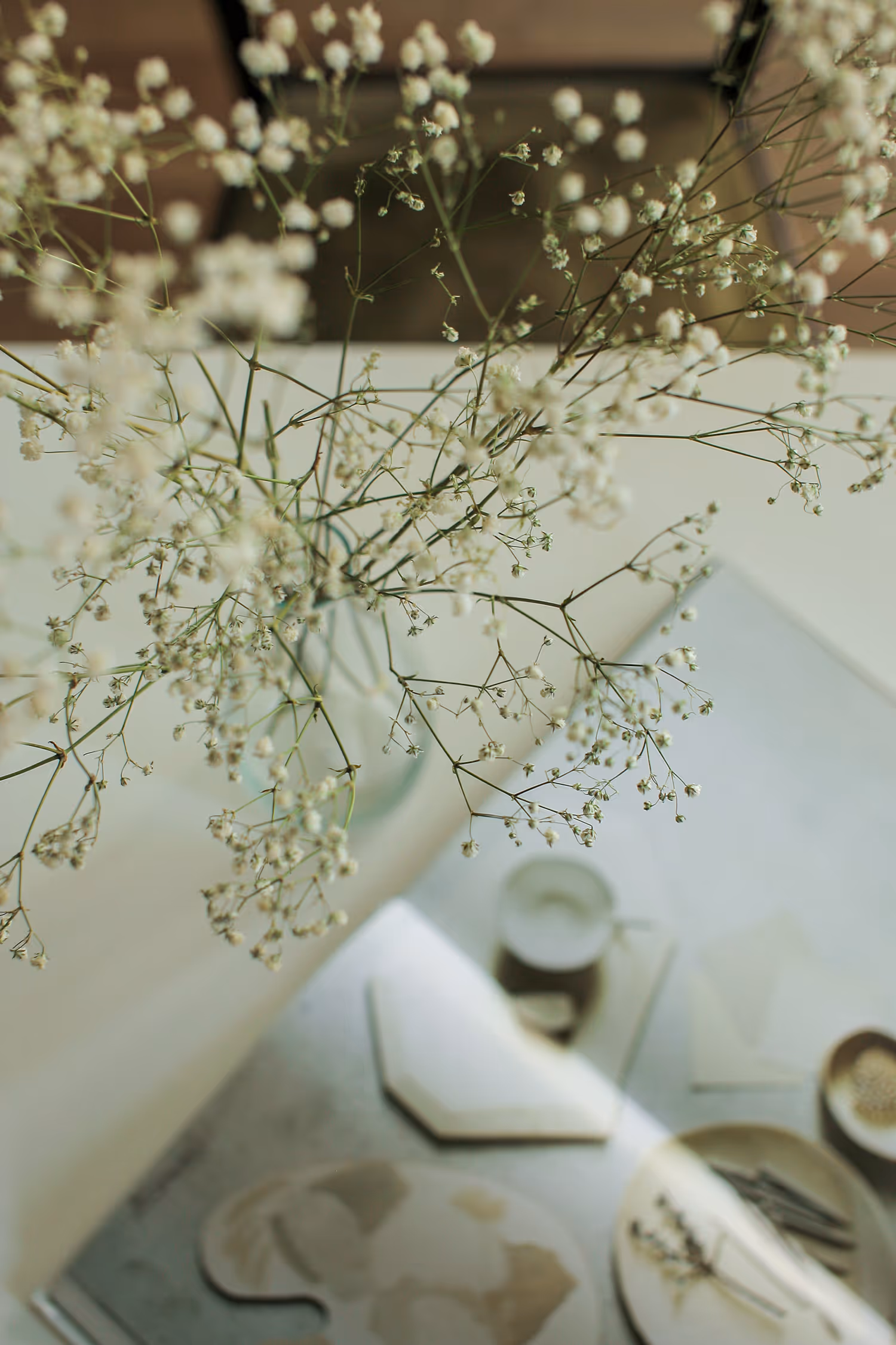 High-key, shallow depth of field lifestyle image of delicate white Gypsophila (Baby's Breath) flowers in a vase on a light-filled table with ceramics and stationery, emphasizing a soft, airy atmosphere.