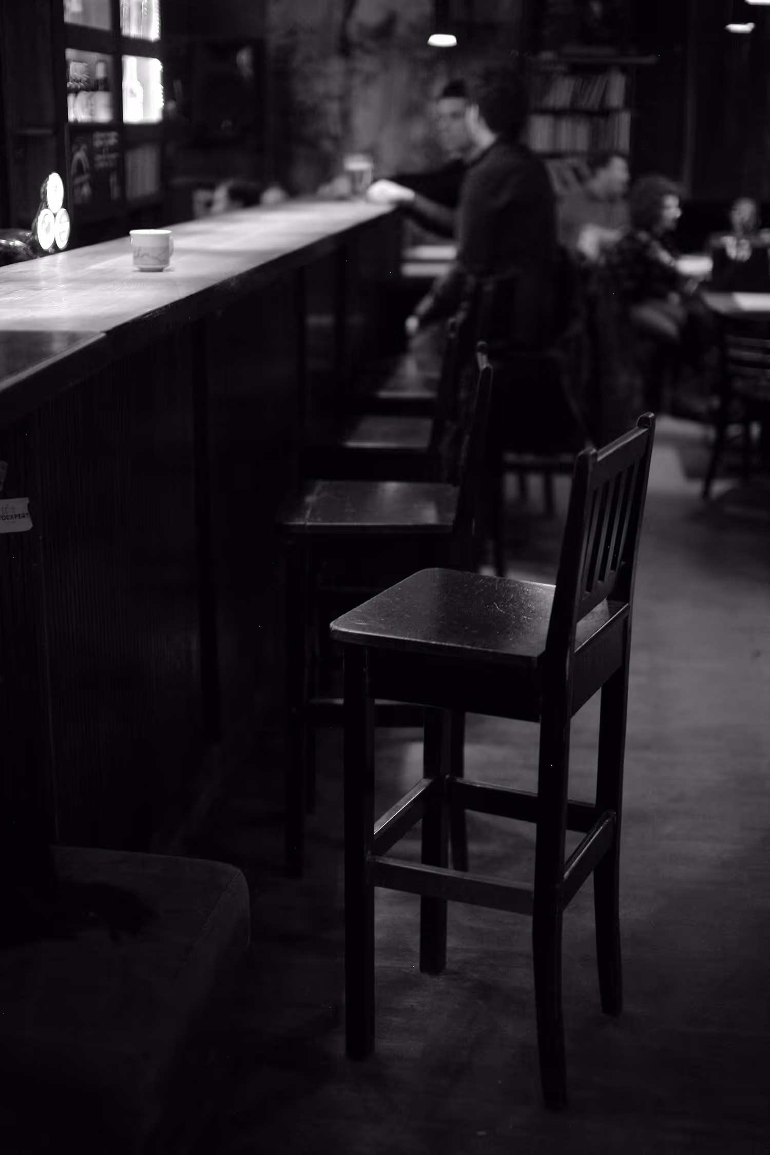 High-contrast monochrome photograph focusing on two empty, tall wooden bar stools in the foreground, with indistinct figures seated at the long bar in the softly lit, dark interior of a cafe, emphasizing leading lines.