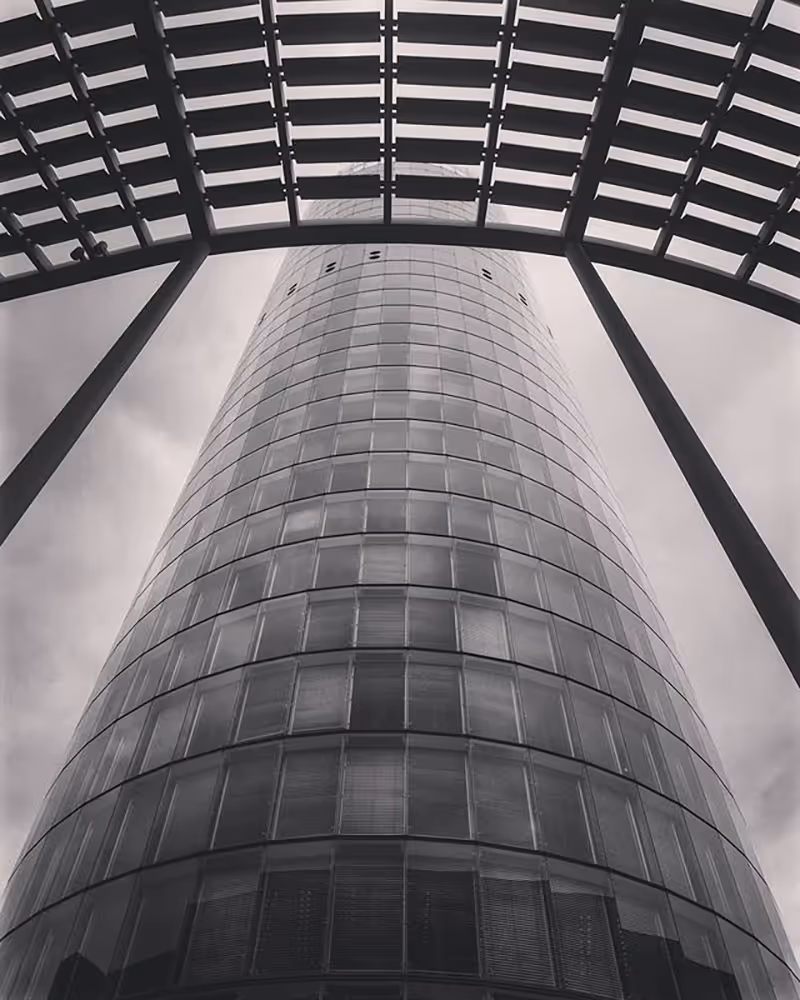 Black and white low-angle, geometric view of the cylindrical RWE Tower in Essen, framed by a dark, angular, slatted structure in the immediate foreground, emphasizing symmetry and the building's height.