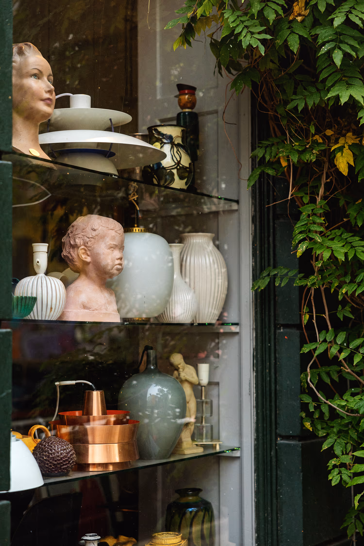 A close-up of a window display in Copenhagen features vintage Scandinavian ceramics, copper cookware, and plaster busts, framed by dark green ivy, emphasizing texture and interior design.