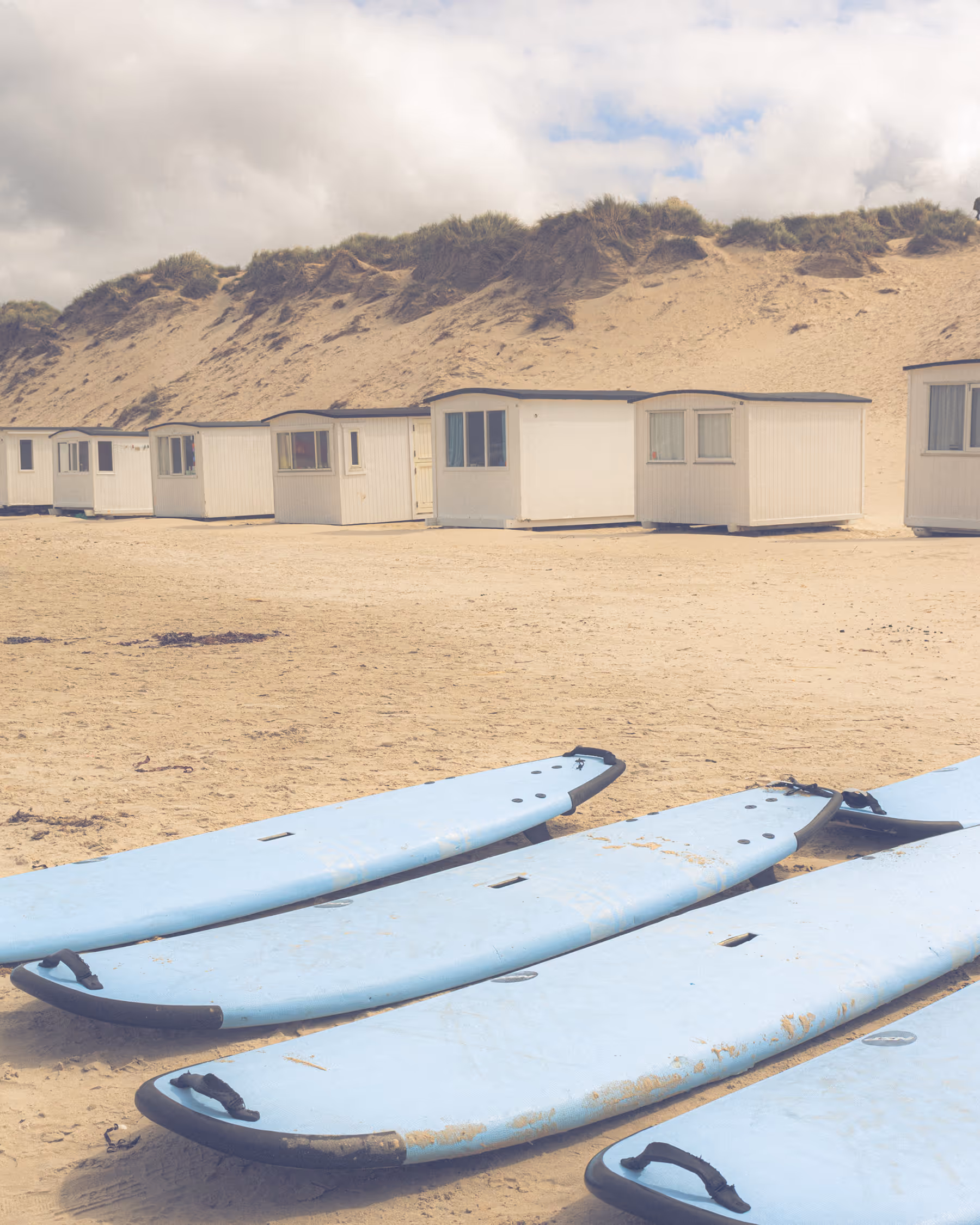 Rows of light blue surfboards lie on the sandy Løkken beach in Denmark, foregrounding minimalist white beach cabins and marram grass-covered dunes under a pale sky.