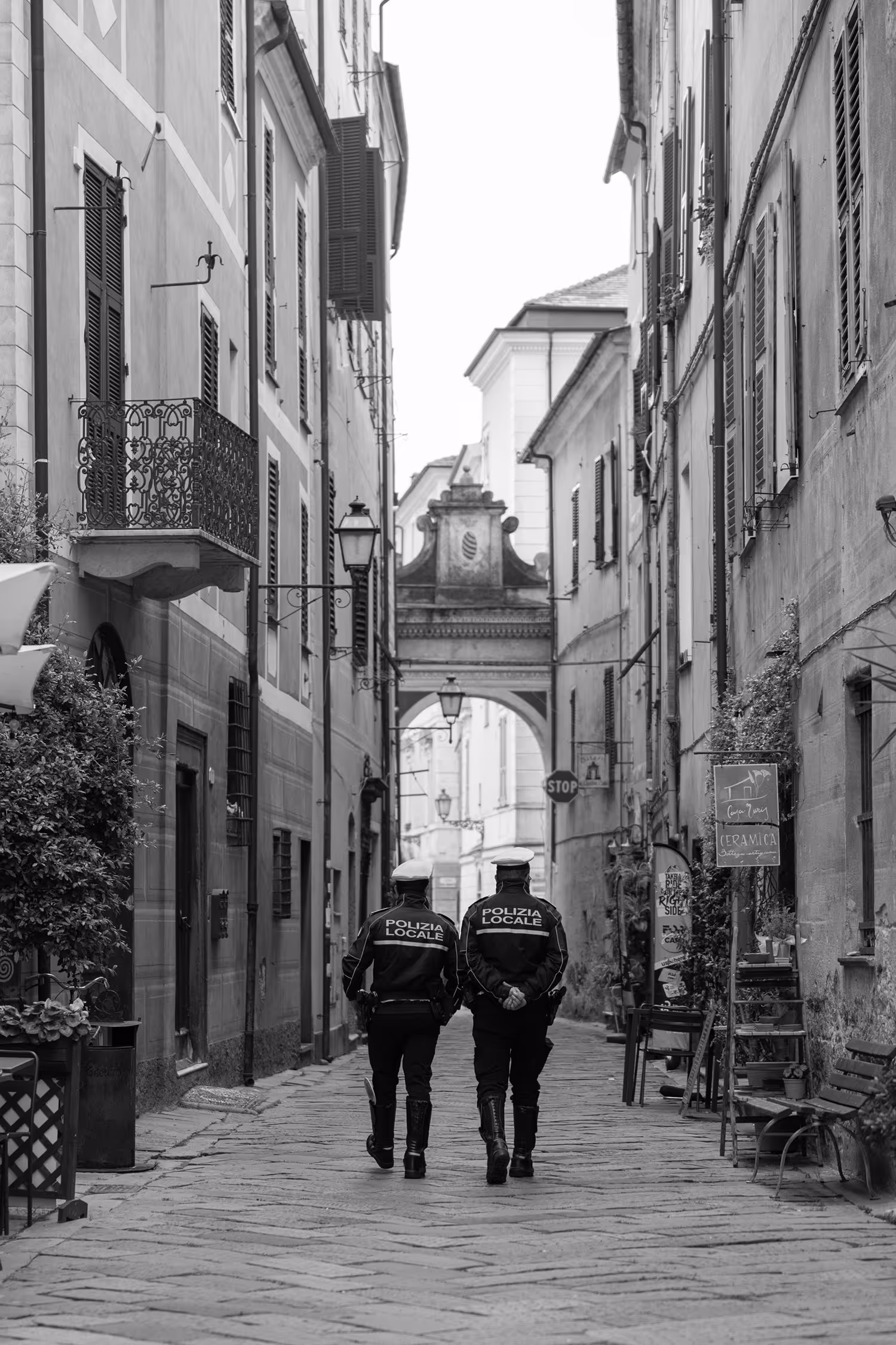 Black and white candid street scene in Finalborgo, Finale Ligure, Italy, showing two "Polizia Locale" officers walking away down a narrow, historic cobblestone alley lined with traditional buildings, framed by an archway in the distance.