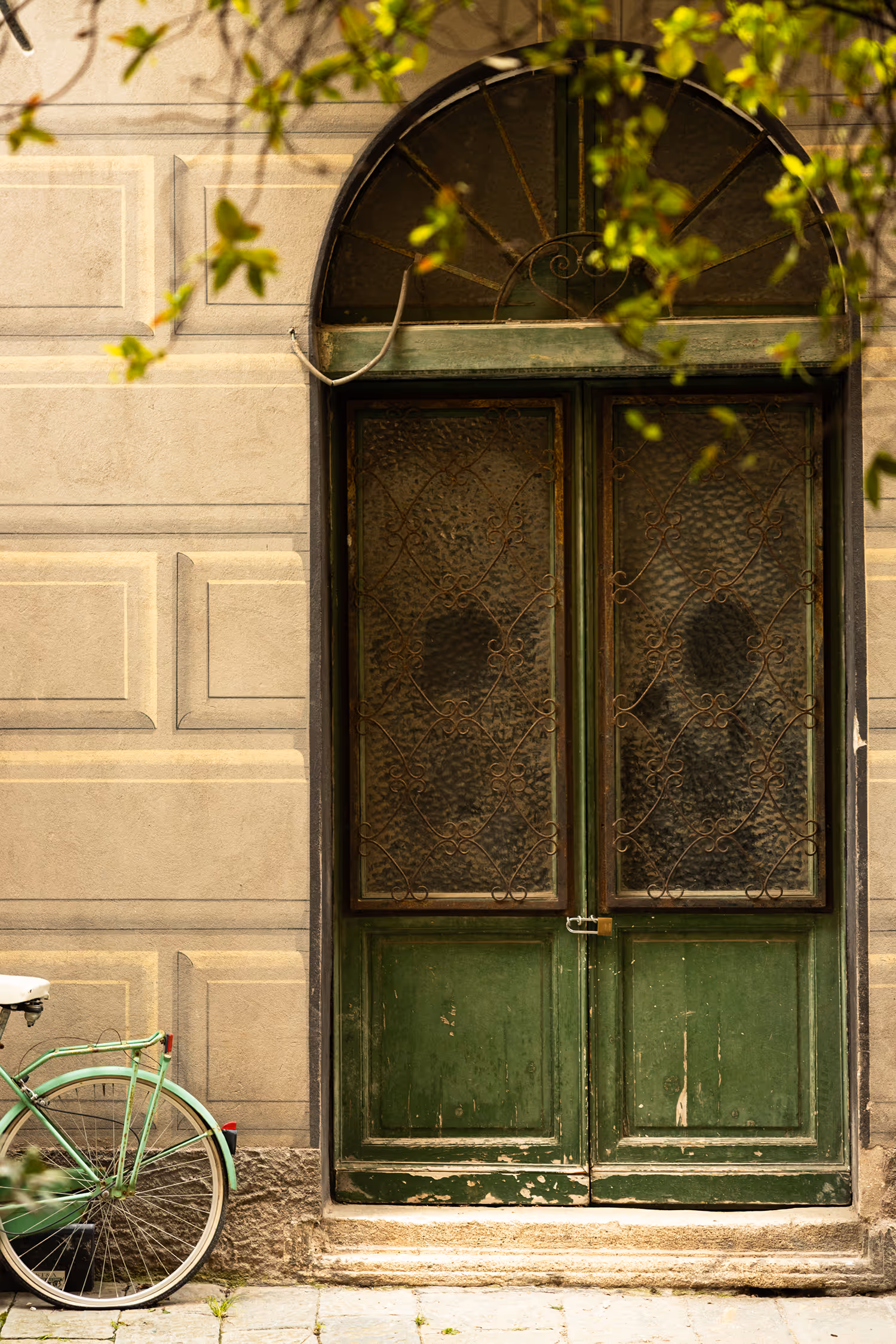 Candid travel photography of a weathered, arched green wooden door in Finalborgo, Finale Ligure, Italy, set against a stone wall, with a vintage mint-green bicycle leaning nearby under sun-drenched foliage.