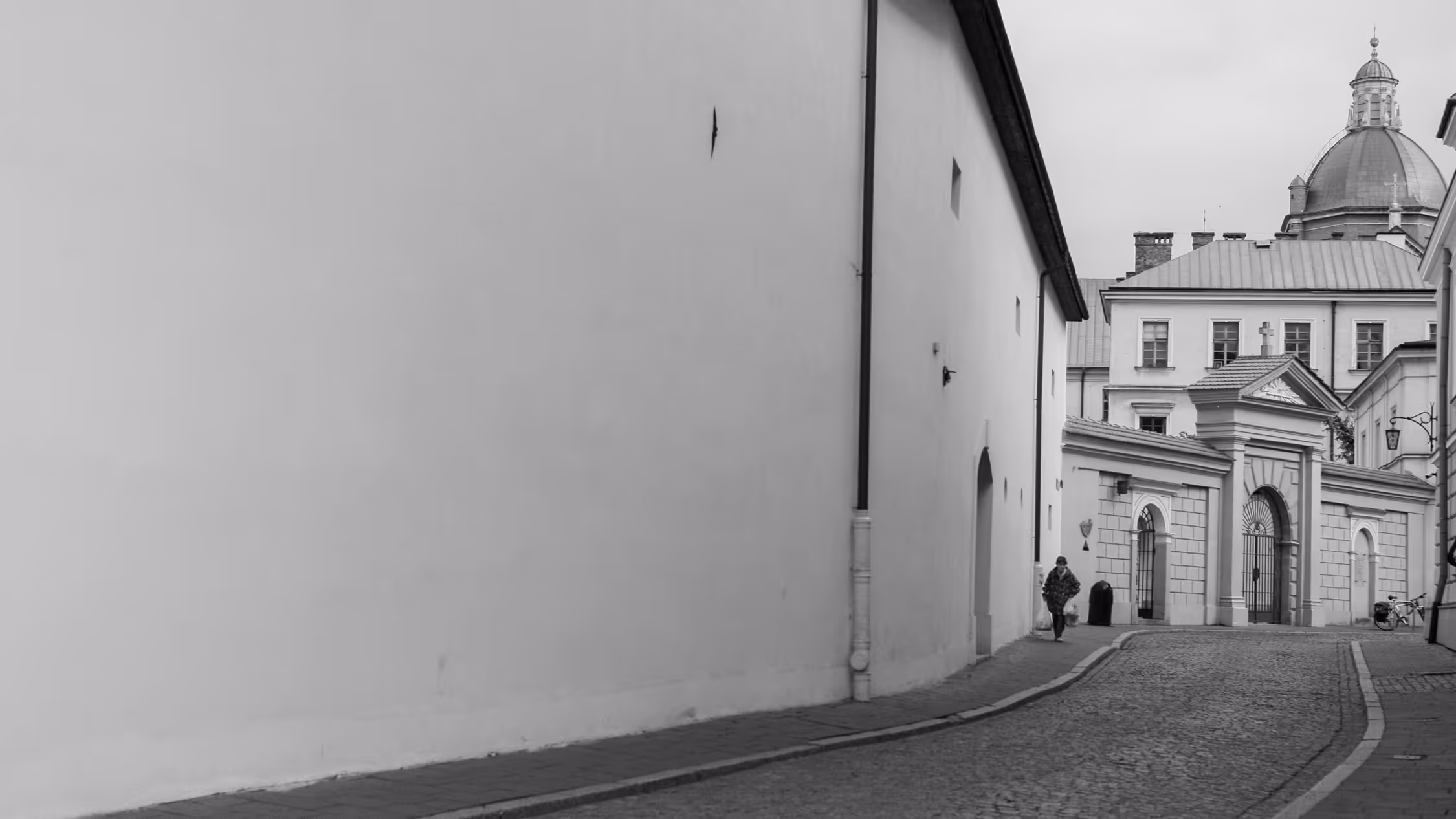 Black and white candid street scene in Kraków, Poland, showing a minimalist view of a massive white wall leading towards the baroque dome and architecture of the Saints Peter and Paul Church, emphasizing negative space and historic urban context.