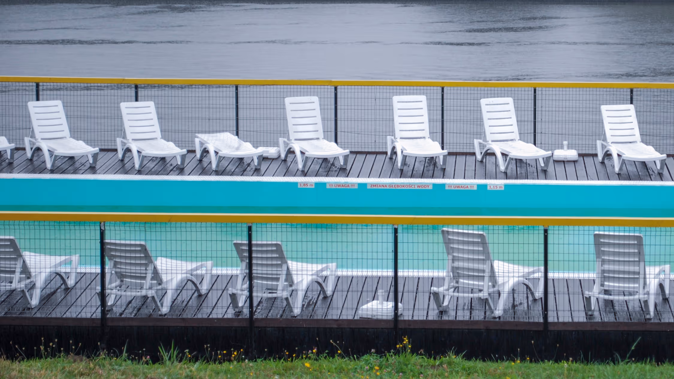 Symmetrical, minimalist composition of empty white sunbeds around a swimming pool under a grey, atmospheric sky, emphasizing repetition and negative space.