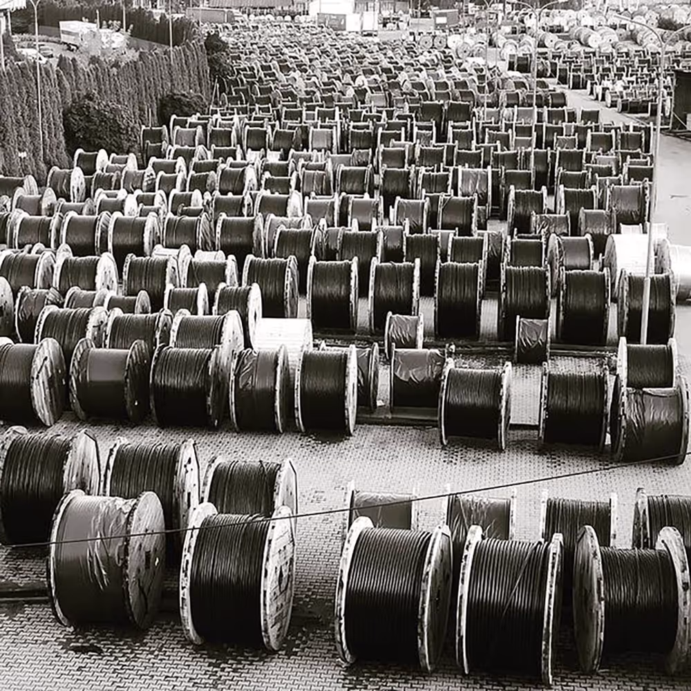 High-angle monochrome photograph of an expansive industrial storage yard in Vienna, filled with hundreds of identical spools of dark cable creating a strong pattern and repetition.