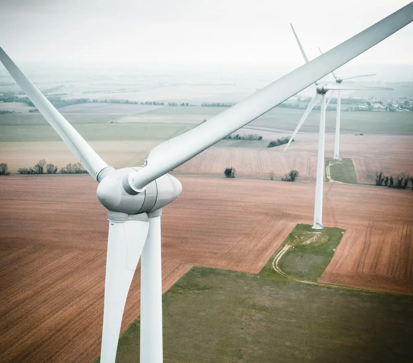 Aerial view of wind turbines standing in farmland, with two turbines close-up in the foreground and patchwork fields stretching to the horizon.