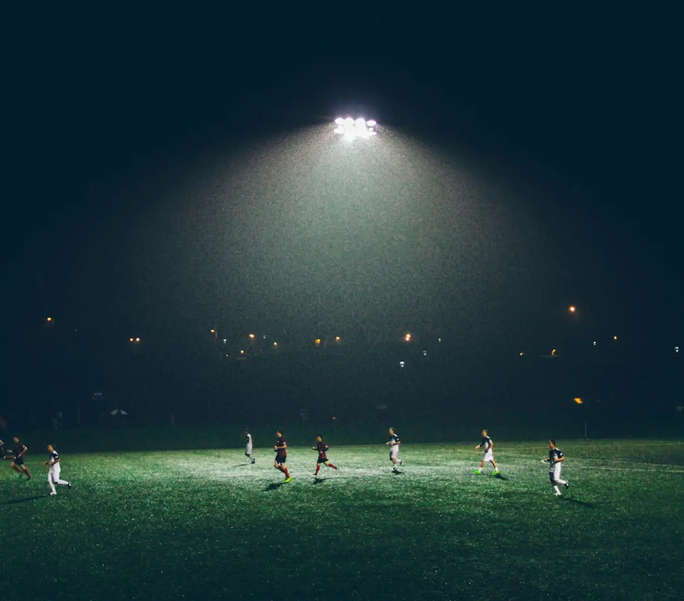 Footballers competing on a floodlit pitch at night, with a single stadium light illuminating the field against a dark sky.