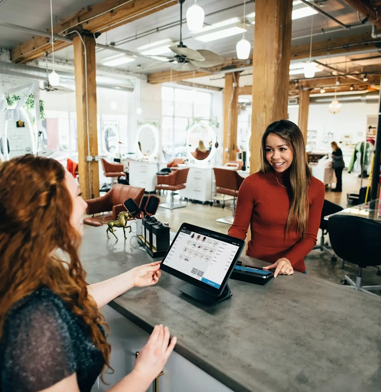 Salon receptionist checking a customer out using a tablet point-of-sale system at the front desk of a bright, industrial-style hair salon.