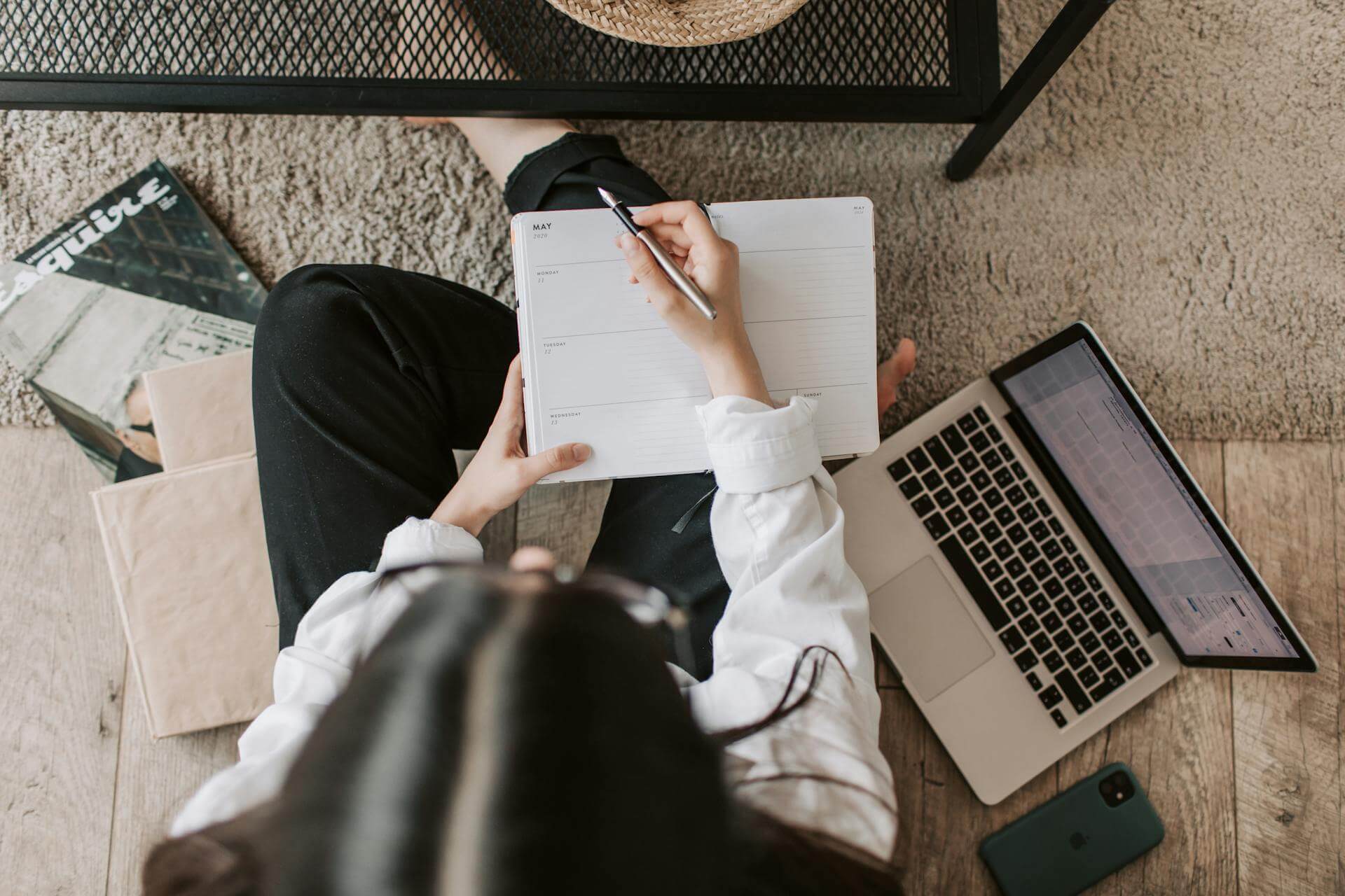 Person in white shirt sitting cross-legged on wooden floor, writing in a planner with a pen, surrounded by laptop, phone, and scattered papers.