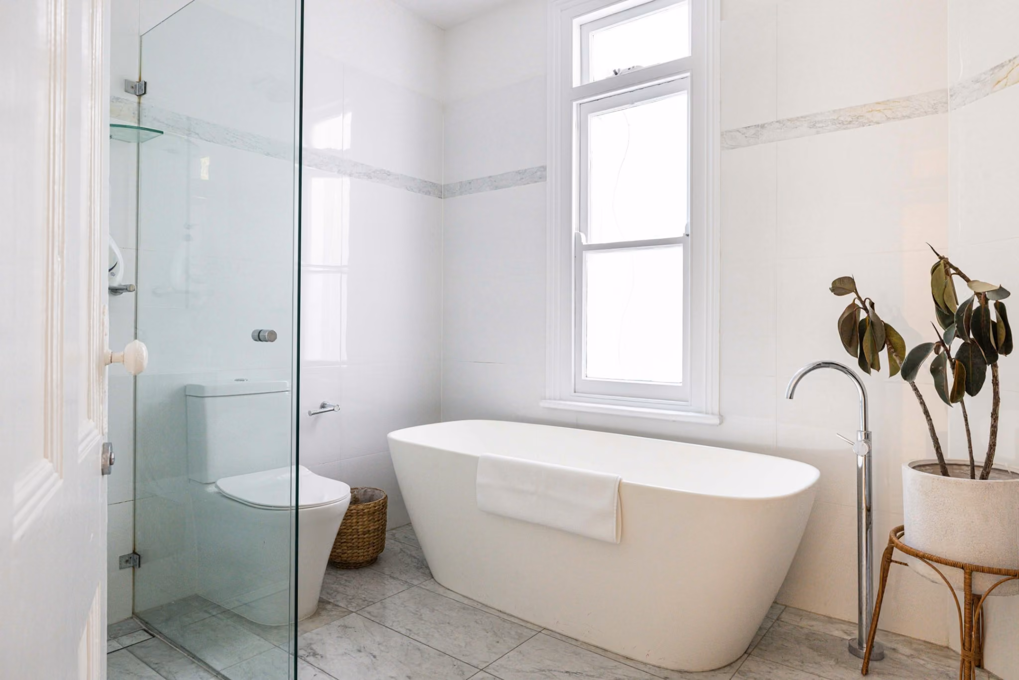 Light-filled bathroom with freestanding bath, marble flooring and restored heritage window.