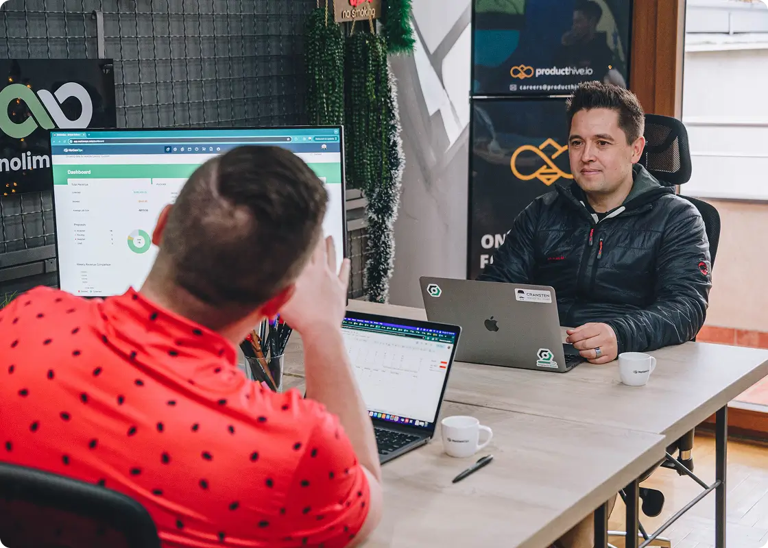 Trevor Maddux in the office, sitting at his computer, with one more person in the image with his back turned