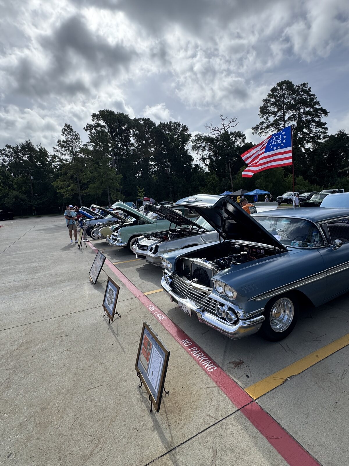 A row of restored classic cars lines the curb beneath an American flag on a summer afternoon at the Golden Corral in Conroe