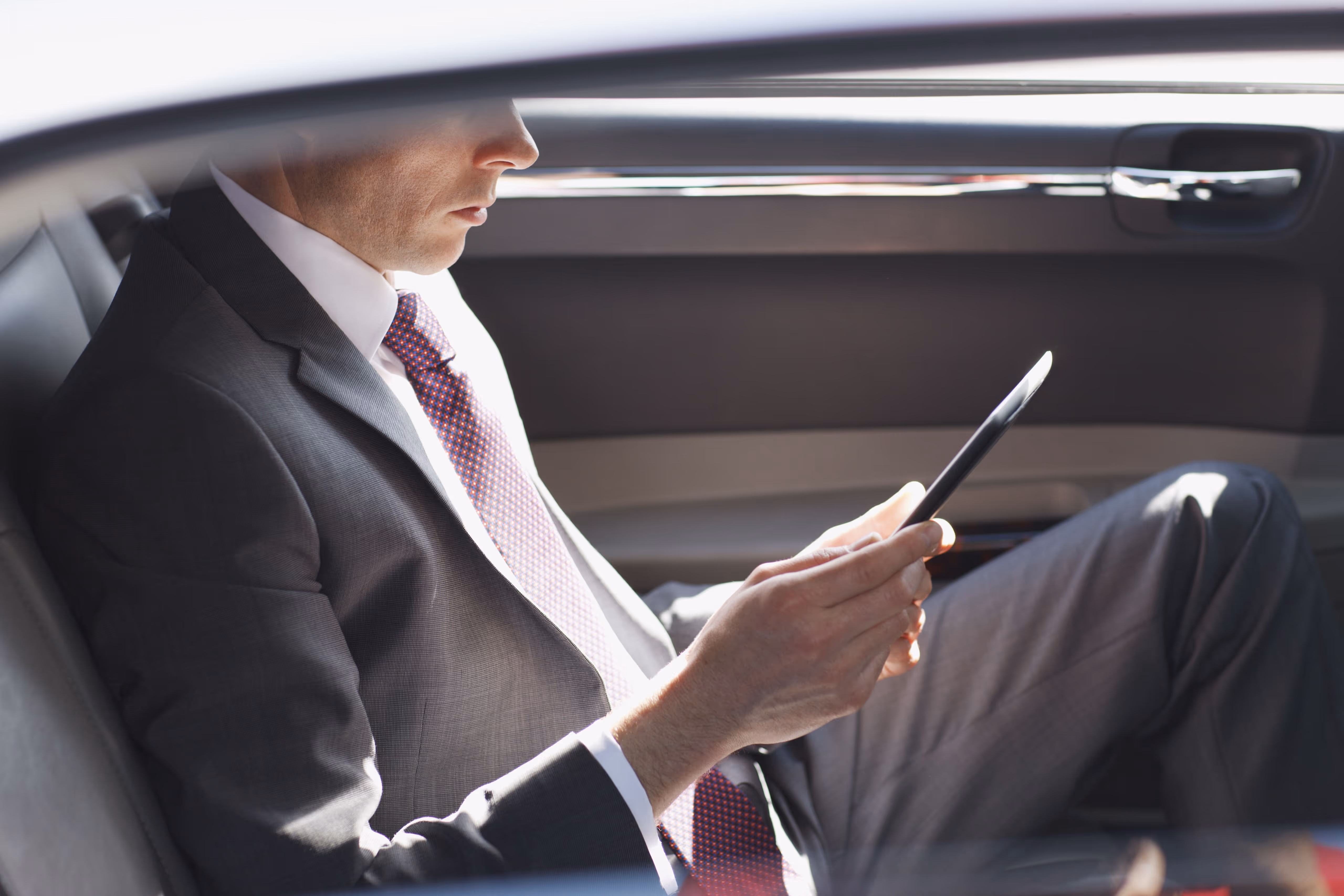 Man in a suit sitting in the backseat of a car looking at a tablet.